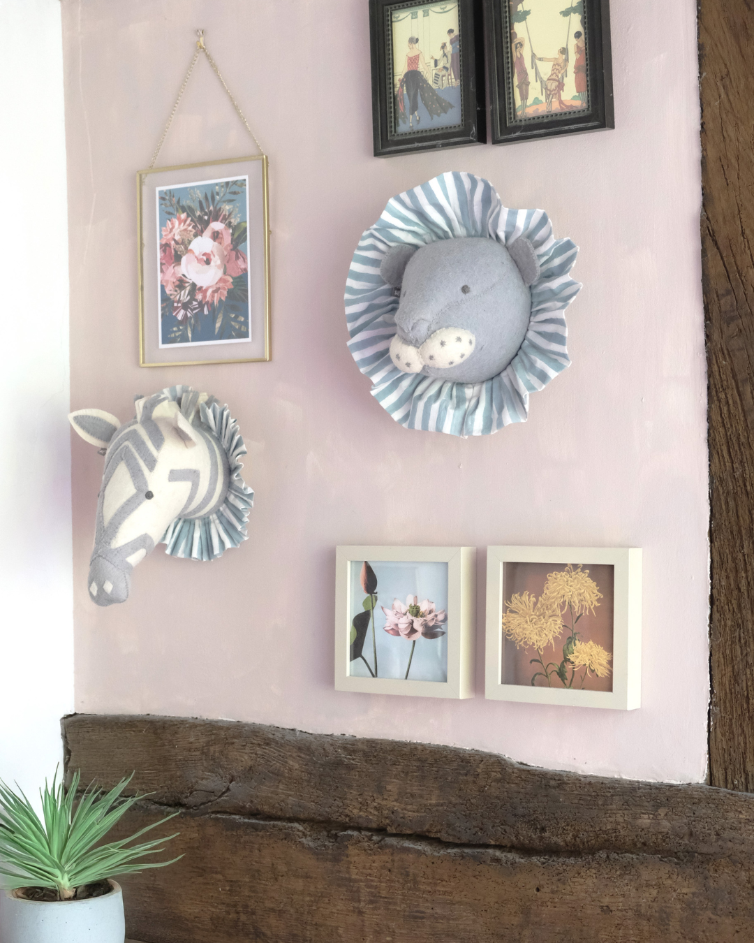 Decorative wall with two framed photos, plush animal wall hangings of a lion and zebra with ruffled manes, and botanical artwork, adorned on a pink wall with a wooden beam at the bottom and a small plant in a pot.