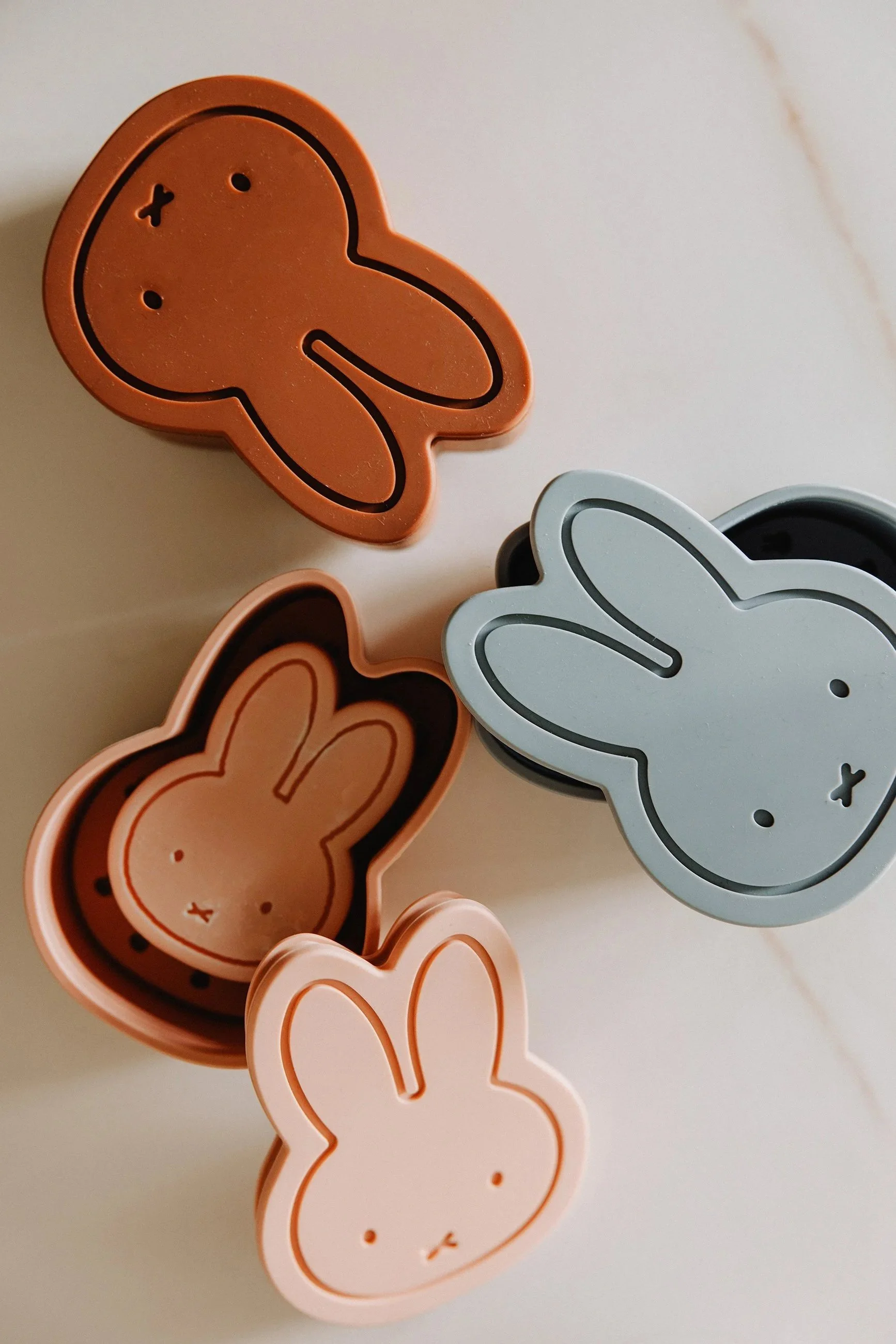 Four bunny-shaped cookie cutters in pastel pink, white, brown, and light blue colors on a light-colored surface.