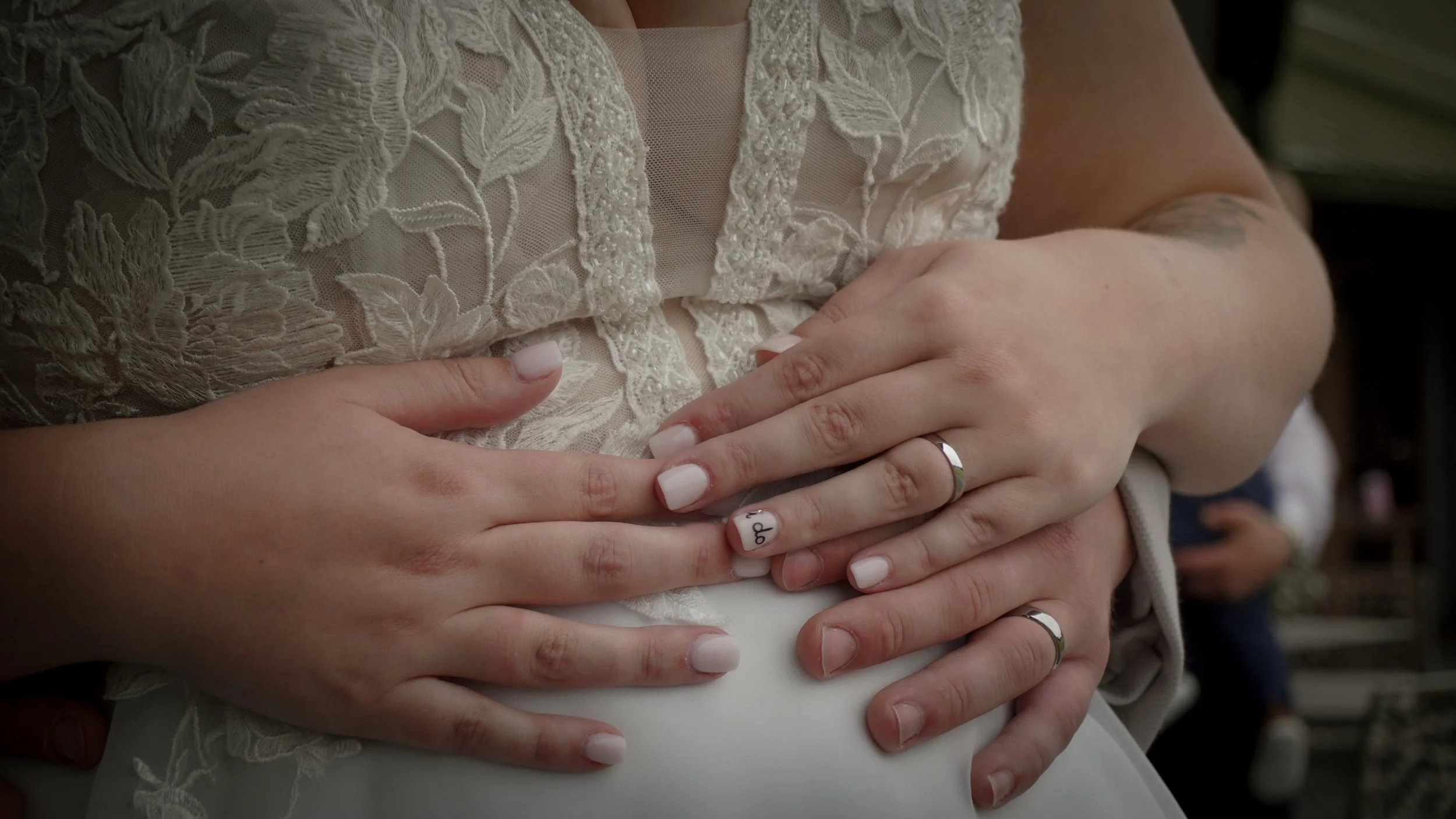 Close-up of a bride and groom holding hands, showing wedding rings on their fingers. The bride is wearing a lace dress and has a manicure with one nail lettered 'Q' and a tattoo partially visible on her arm in the background.