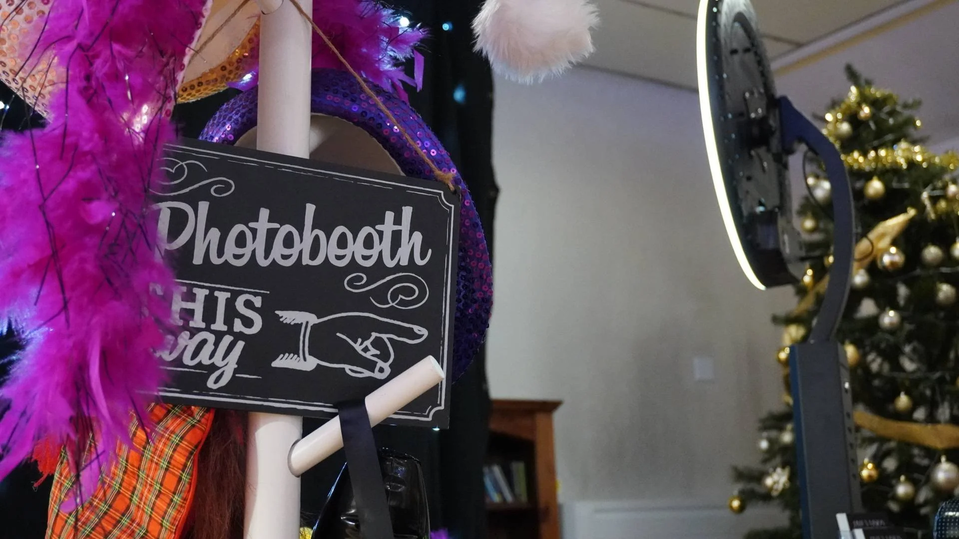A photo booth setup with a sign that reads 'Photobooth this way,' decorated with pink feathers, purple sequin trim, and a white pointing hand sculpture. In the background, a Christmas tree decorated with gold ornaments is visible.