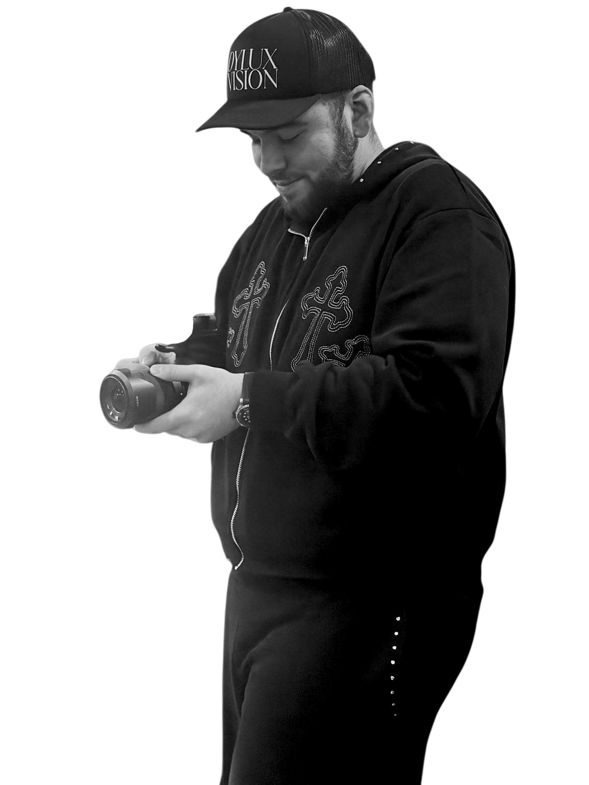 A man in a black baseball cap with the phrase 'DYLUX VISION' on it, wearing a black jacket with decorative embroidery, holding a camera and looking down at it.
