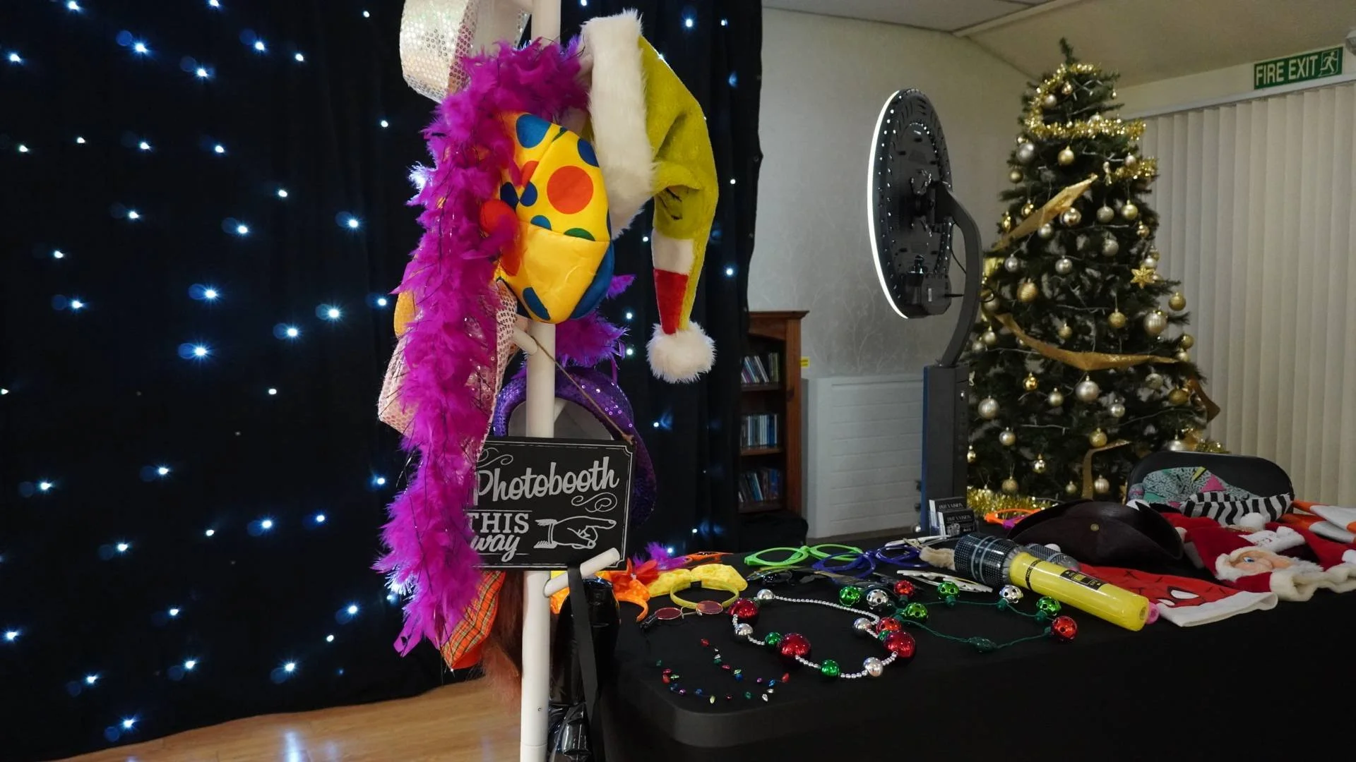 Table with various Christmas-themed items in a photo booth, including hats, necklaces, sunglasses, and costumes, in front of a decorated Christmas tree.