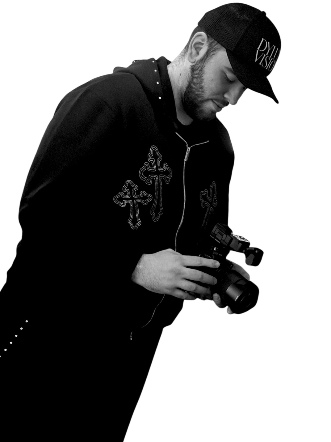 A young man wearing a black baseball cap, black hoodie, and black jacket with cross designs, holding a camera, and looking down.