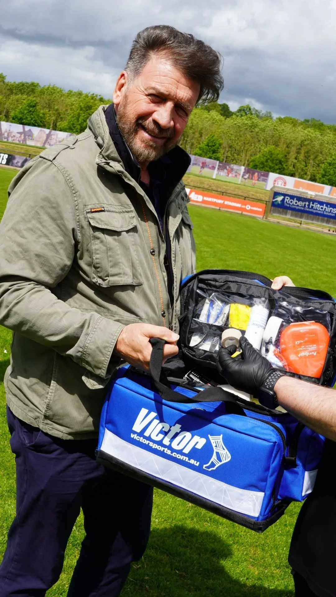A man with gray hair and a beard smiling while holding a medical kit, with another person's hand handing him supplies. They are outdoors on a grassy field on a cloudy day, with trees and advertising boards in the background.
