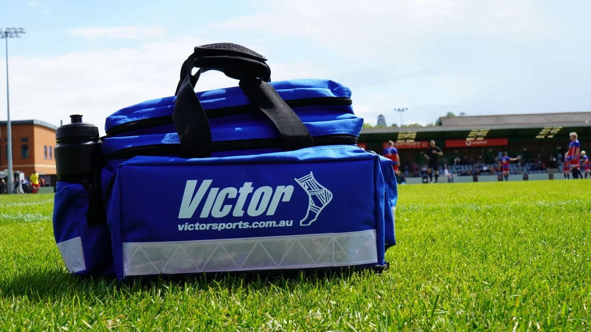 A blue sports bag with Victor branding and a water bottle on a grass field during a cricket game, with players and a stadium in the background.