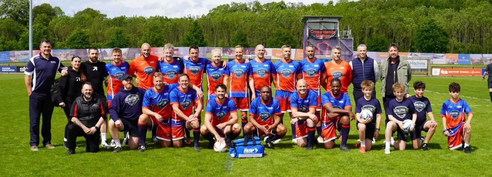 Group of rugby players and officials on a grass field, some holding rugby balls, posing for a team photo with a backdrop of trees and advertisements.