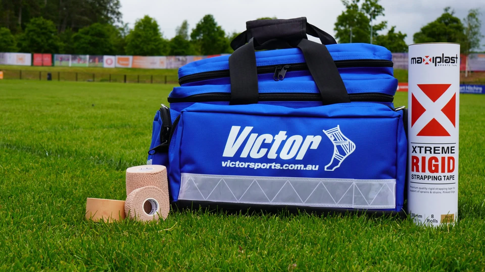 A blue Victor sports bag on grass with a roll of athletic tape, a pink elastic bandage, and a flexible limb support wrap nearby, on a sports field with trees and advertising banners in the background.