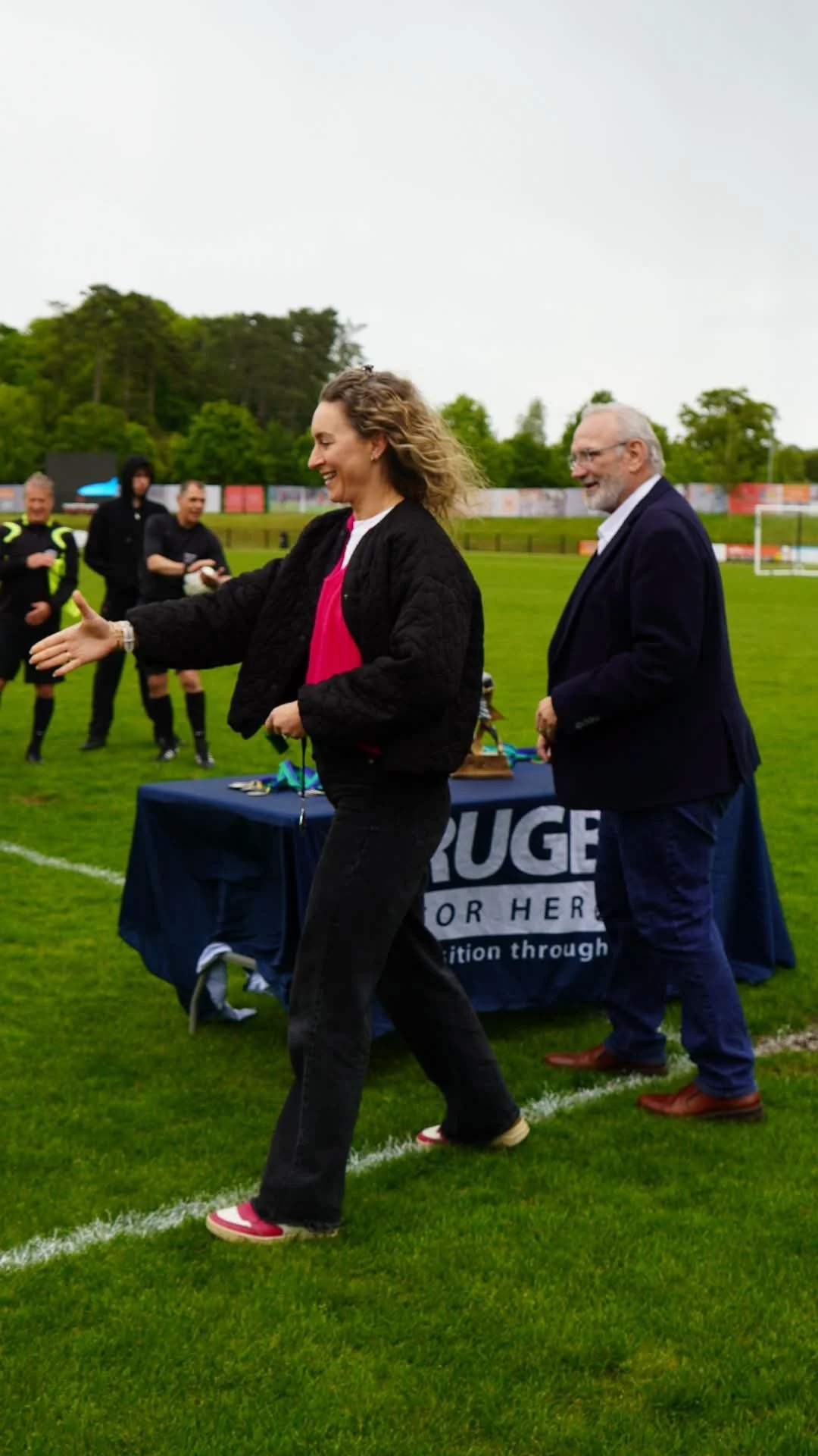Women smiling and shaking hands on a soccer field during an award ceremony. Several people in black sports uniforms are standing in the background. A table with medals and a banner are visible.