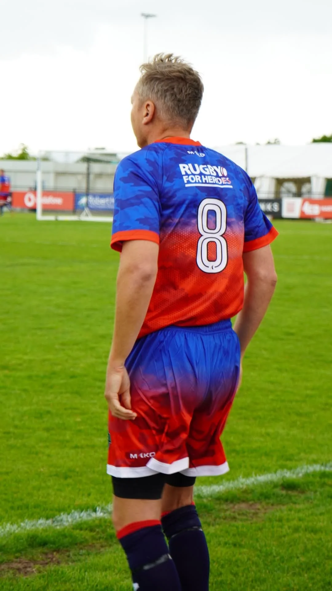 Back view of a male rugby player in a blue and red uniform standing on a grass field with goalposts in the background.
