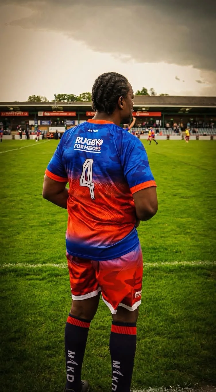 A rugby player in a blue and red jersey with the number 4, standing on a grassy field, looking towards the game under dark, cloudy skies.