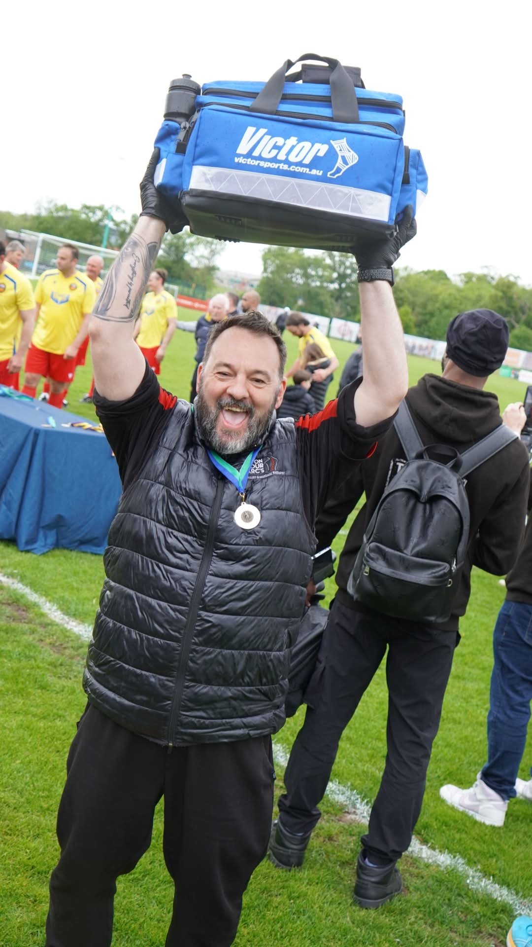 Man with a beard smiling and holding a blue sports bag with the brand Victor, wearing a black vest and black pants, standing on a grassy sports field with people in sportswear in the background.