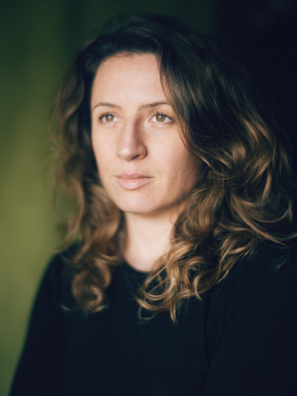 Close-up of a woman with long, curly brown hair and light skin, gazing thoughtfully to the side, wearing a black top, blurred green background.