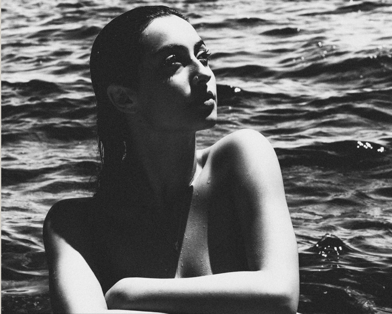 A black and white photo of a woman with wet hair, partially submerged in water, with an intense expression and light falling on her face and shoulder.
