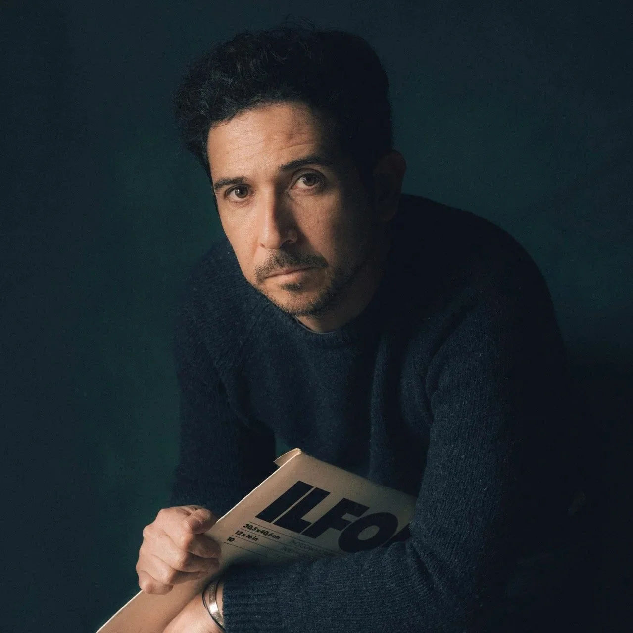A man with dark hair and a beard wearing a dark sweater, holding ILFORD PHOTOGRAPHY SHEETS BOX, looking directly at the camera against a dark background.