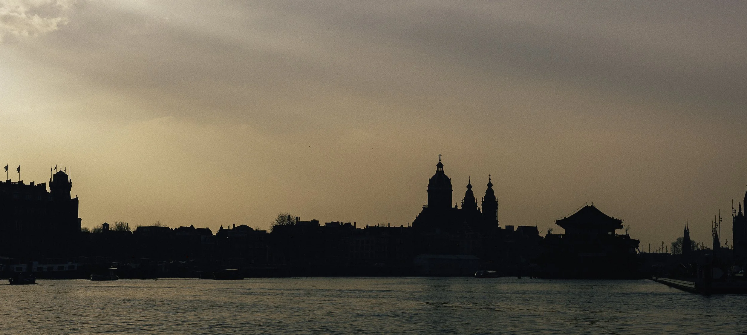 Silhouette of historic buildings and spires along a waterfront at sunset or dawn, with cloudy sky.