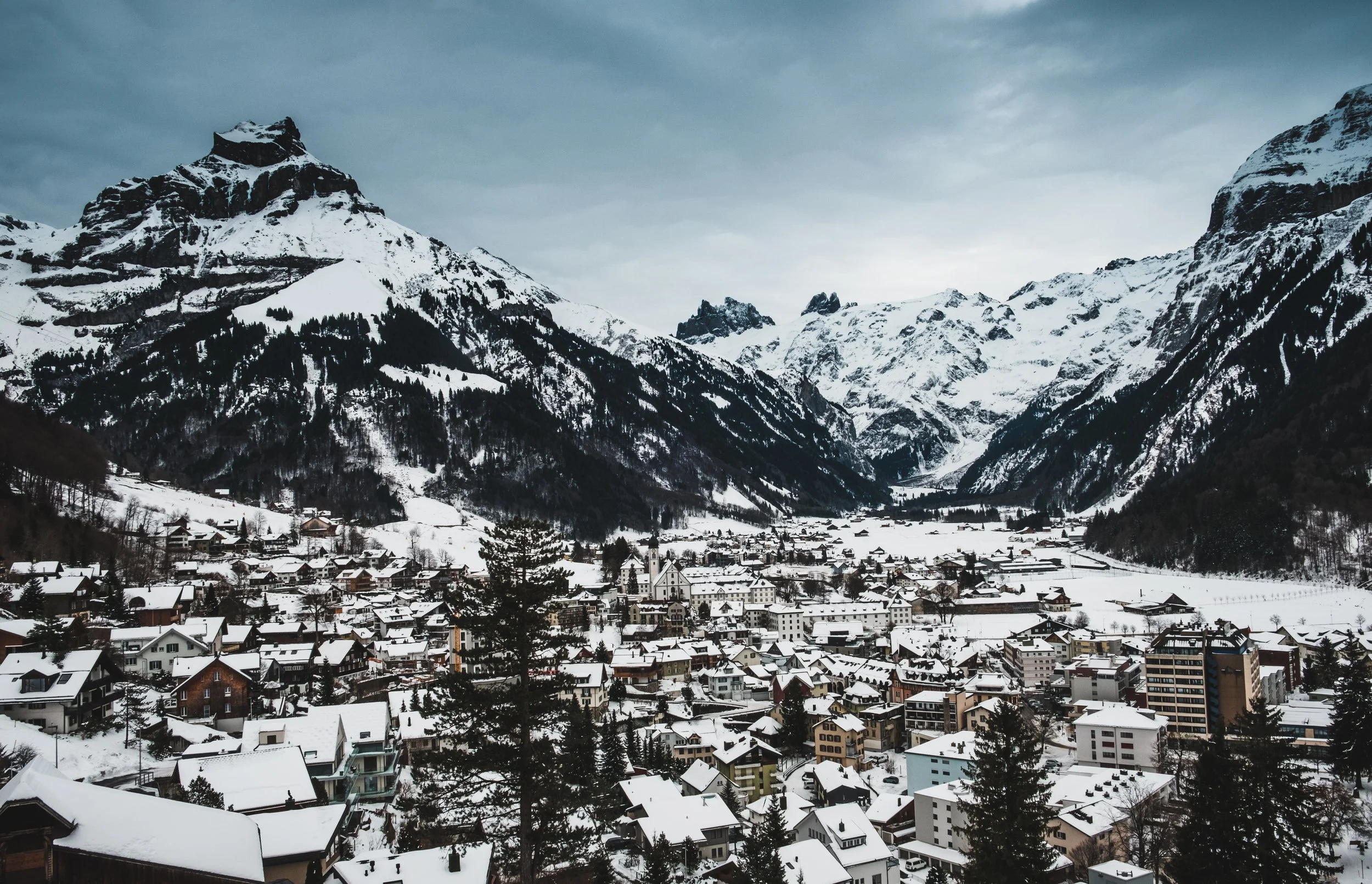 A snowy mountain village with snow-covered roofs set in a valley surrounded by tall, snow-capped mountains on a cloudy day.