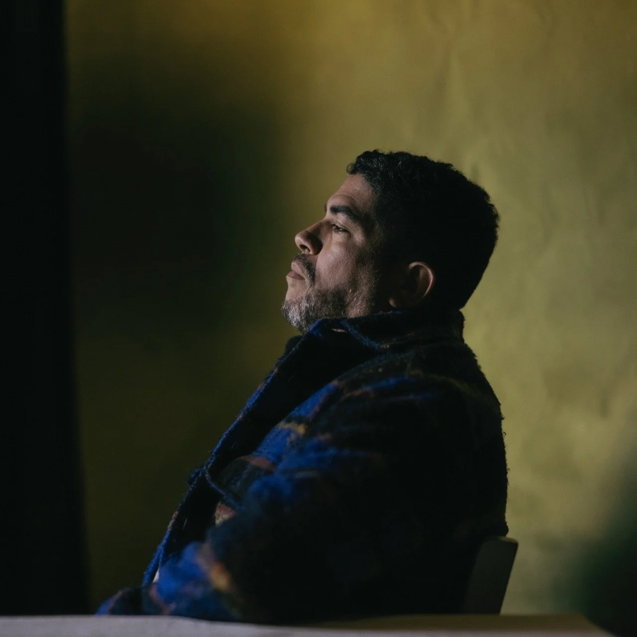 A man with a beard and mustache, sitting with his eyes closed, appears to be relaxed, wearing a colorful, patterned coat against a yellowish background.