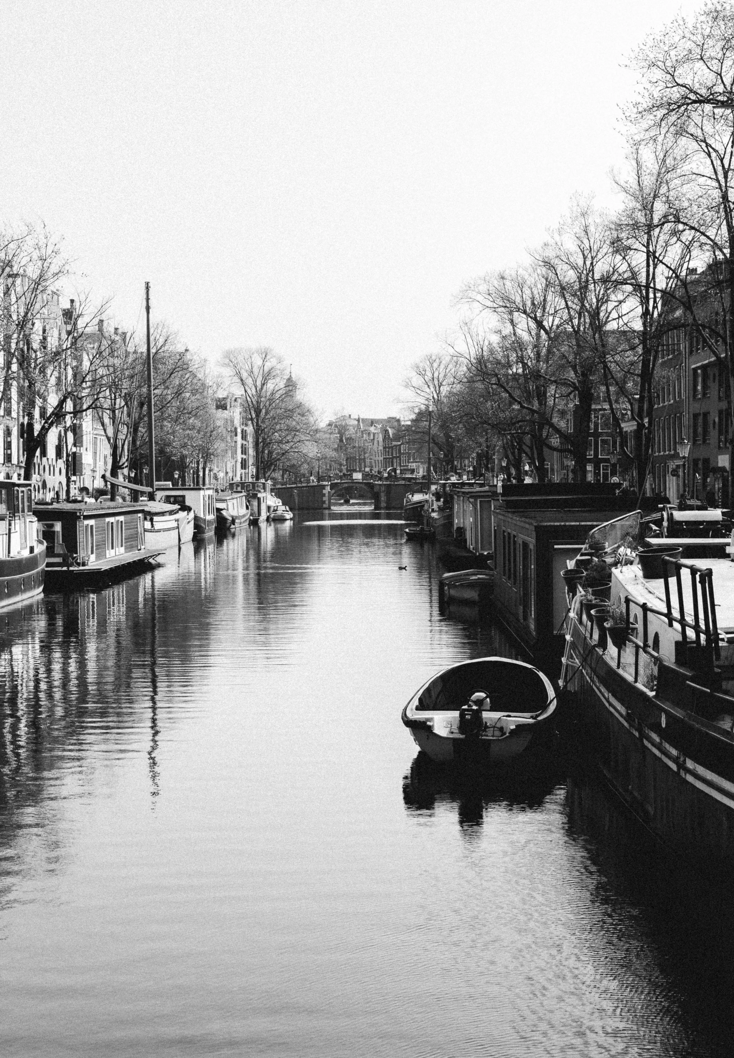 Black and white photo of a canal with boats, surrounded by trees and historic buildings, with a bridge in the background.