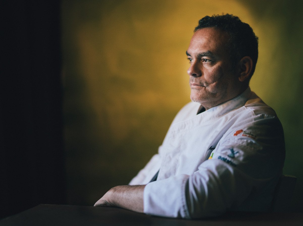 A man in a white chef's coat with colorful patches on the sleeve sits thoughtfully with a serious expression, staring to the side, against a muted yellow background.