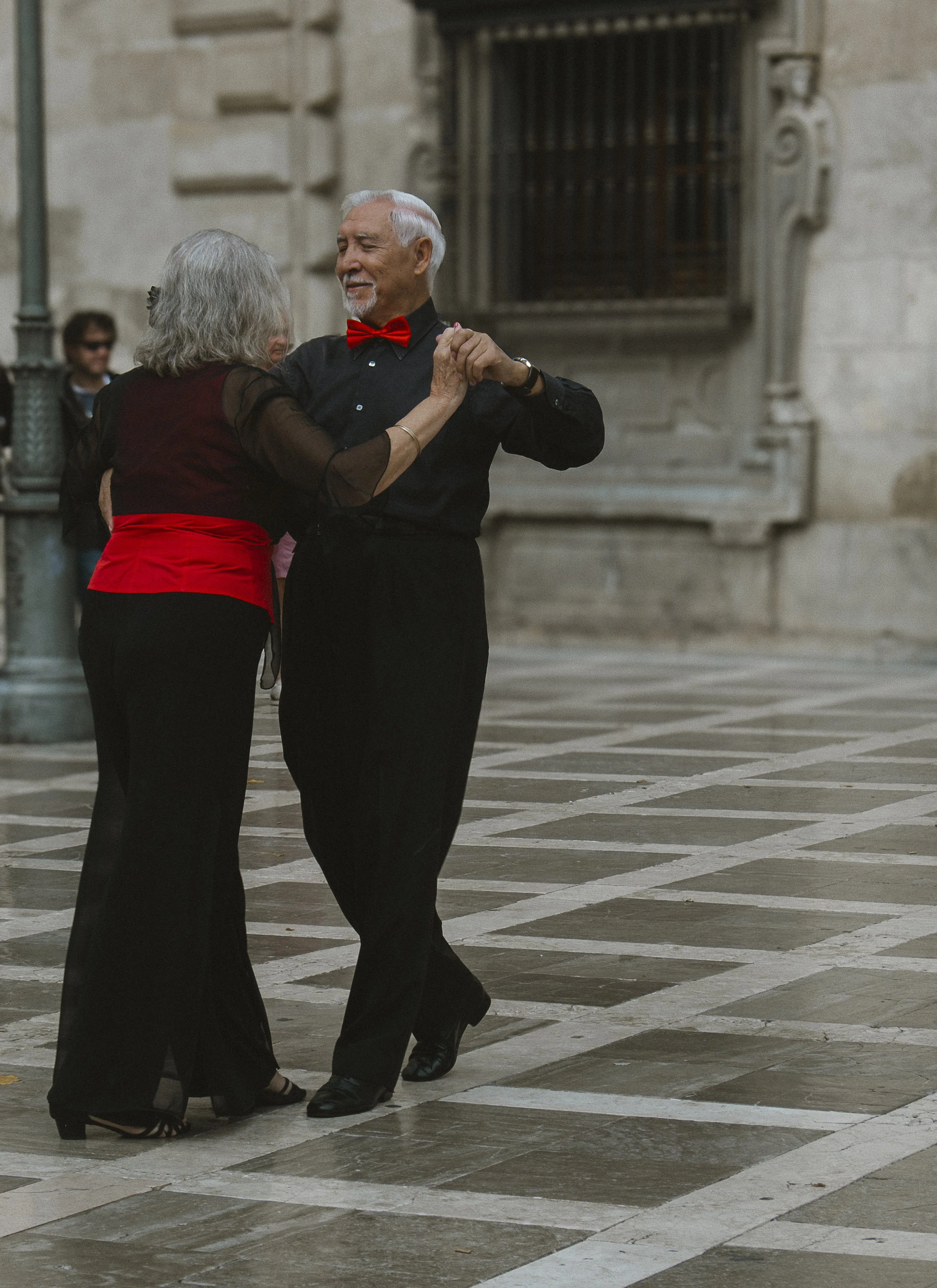 An elderly couple dancing together outdoors on a stone-paved area, with the man holding the woman's hand and the woman looking at him. The man wears a black shirt with a red bow tie and pants, while the woman wears a black dress with a red sash. In t