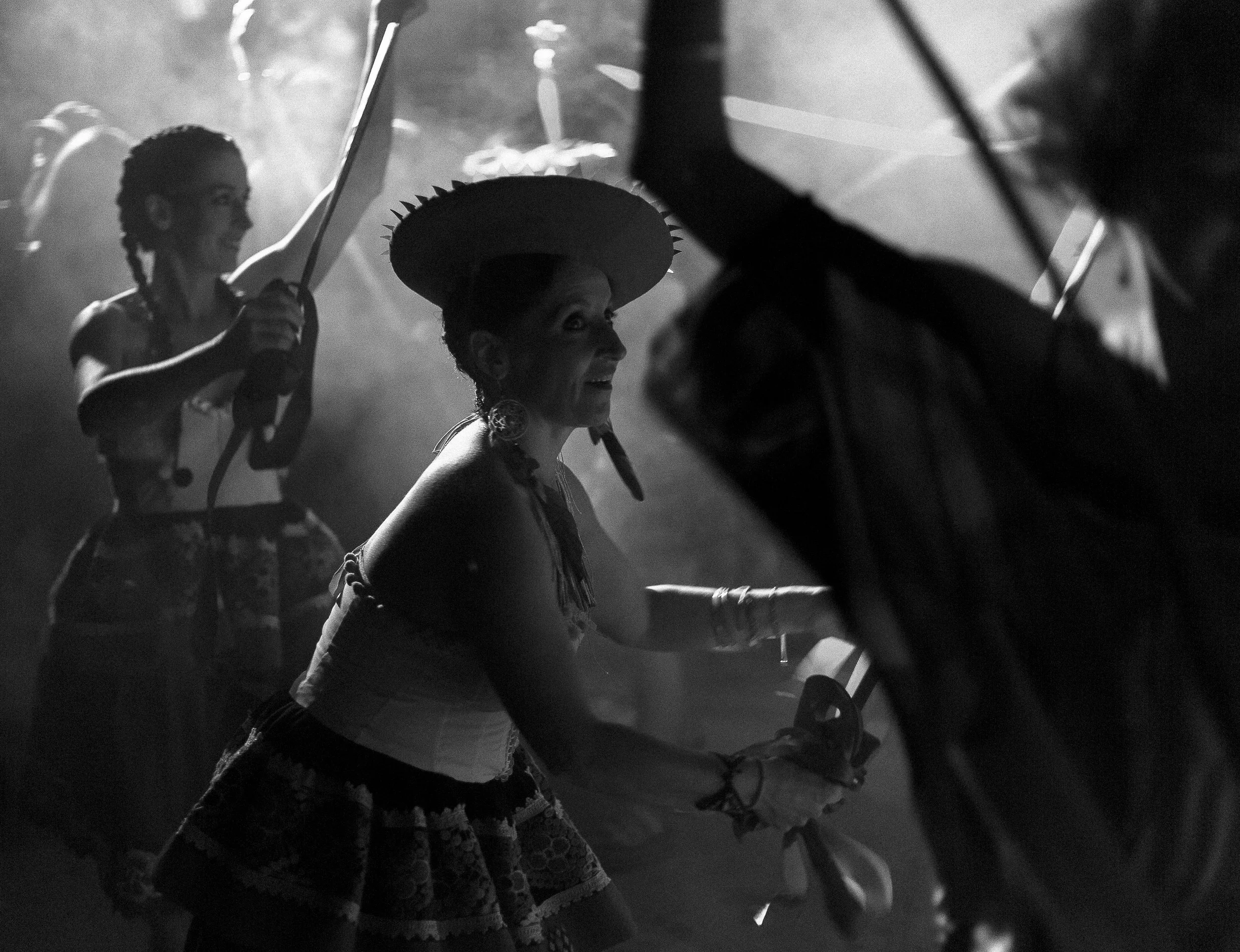 Black and white photo of three women in traditional Mexican attire, holding umbrellas, with one woman wearing a sombrero, in a dance or celebration scene.