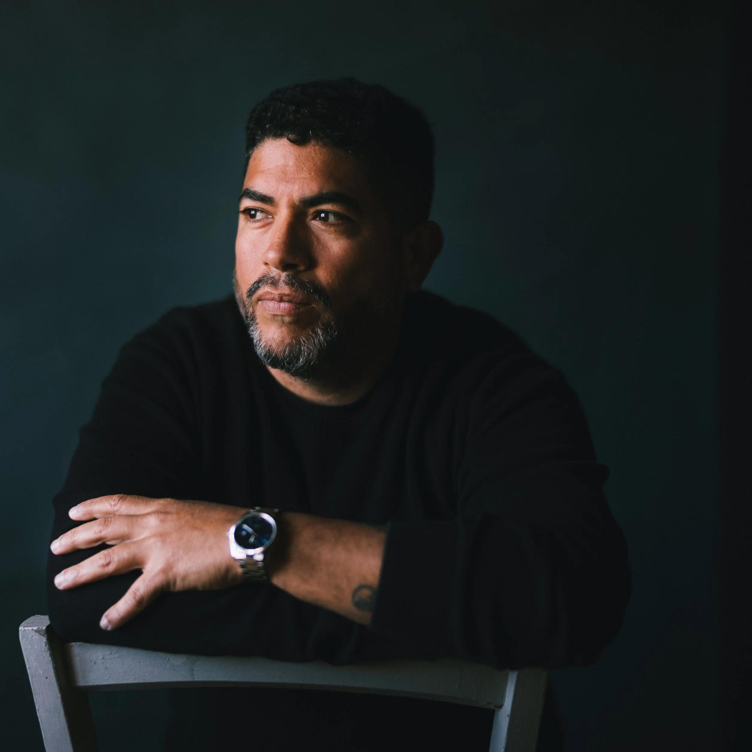 A man with dark hair and a beard looking thoughtfully to the side, wearing a black long-sleeve shirt and a silver watch, sitting with his arms crossed on a gray chair against a dark background.
