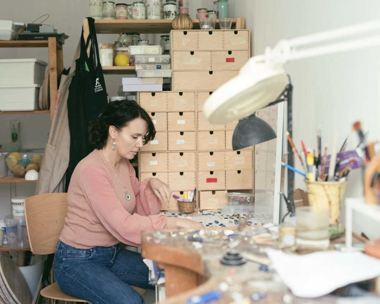 A woman working at a cluttered craft or jewelry-making workspace with organized drawers, tools, and supplies.
