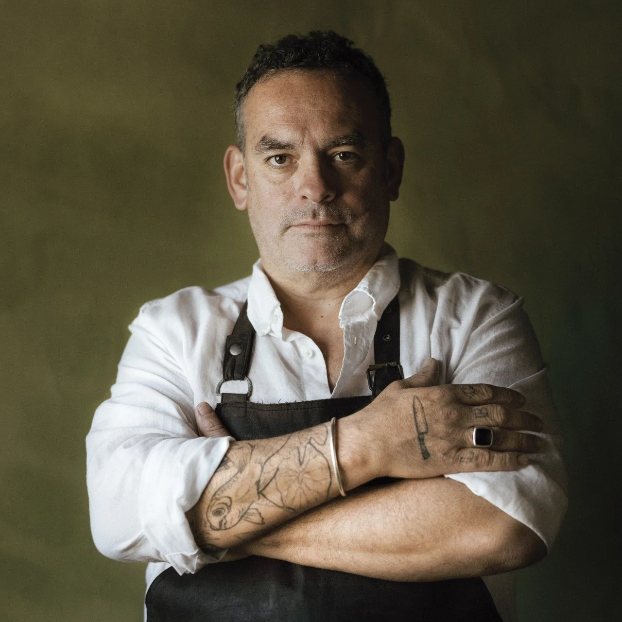 A man with short dark hair and tattoos on his arms, wearing a white shirt and black apron, posing with arms crossed against a dark green background.