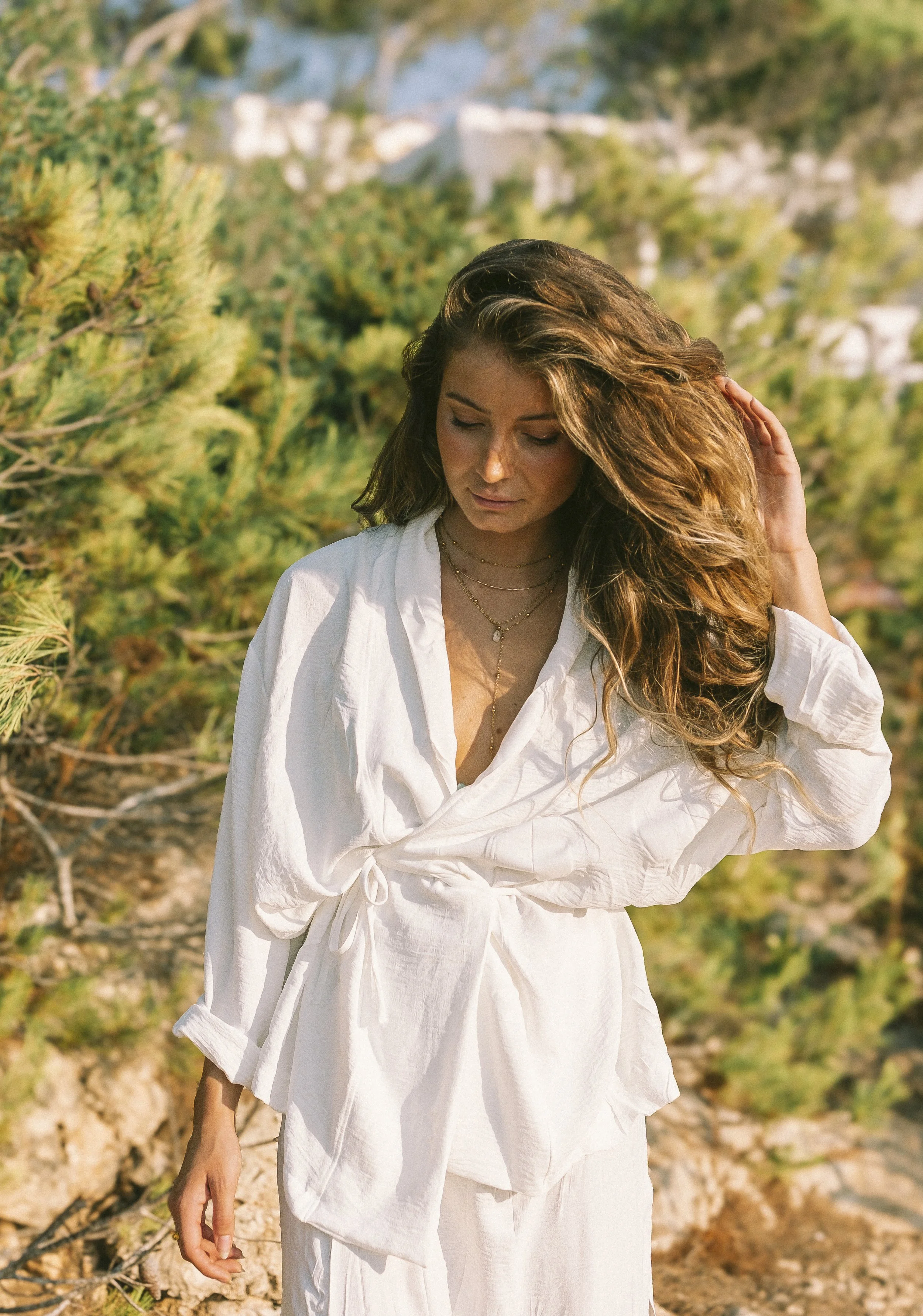 A woman with long wavy hair, dressed in a white shirt and skirt, standing outdoors among green bushes, looking down and touching her hair.