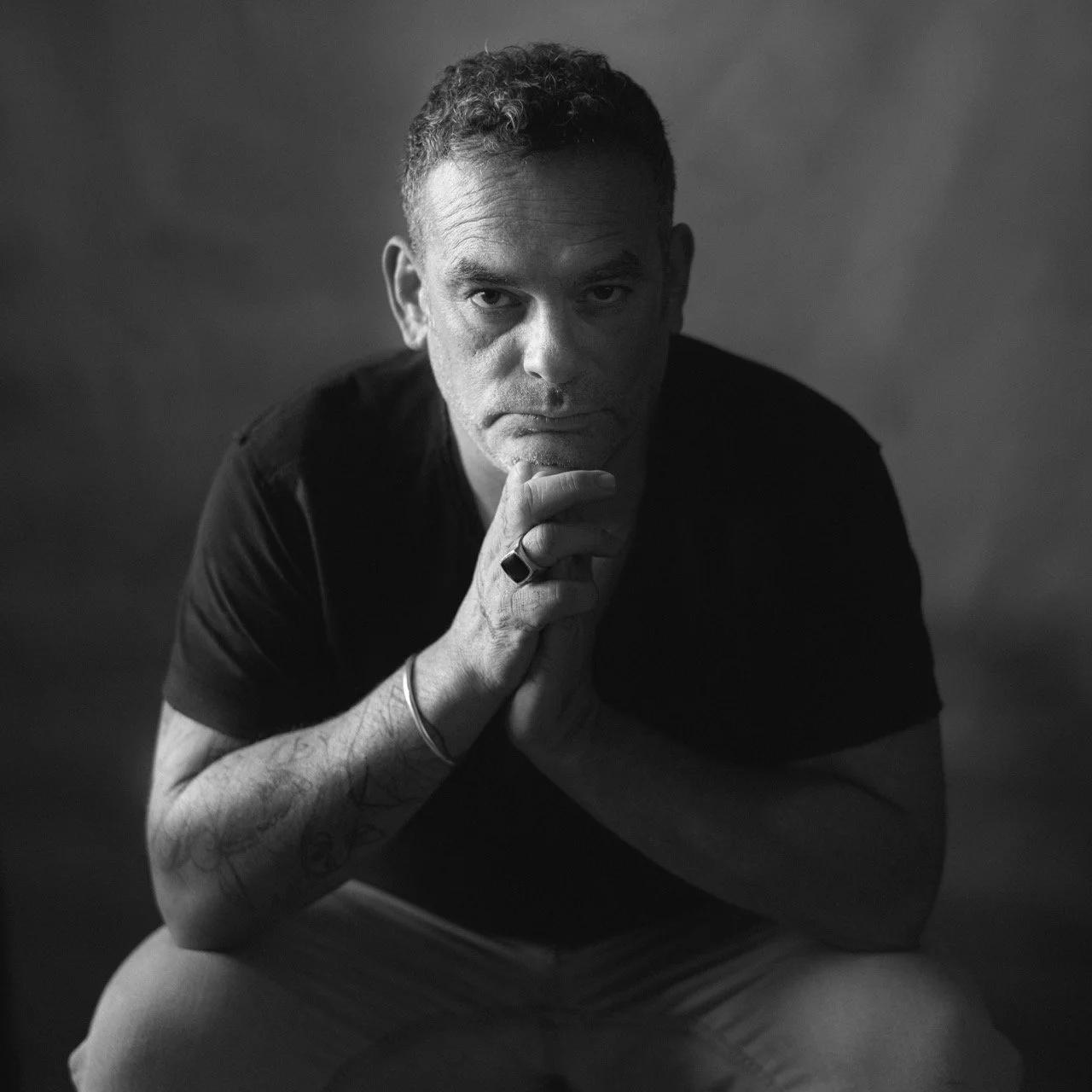 Black and white portrait of a man with short curly hair, wearing a black shirt, sitting with his hands clasped under his chin, looking directly at the camera.