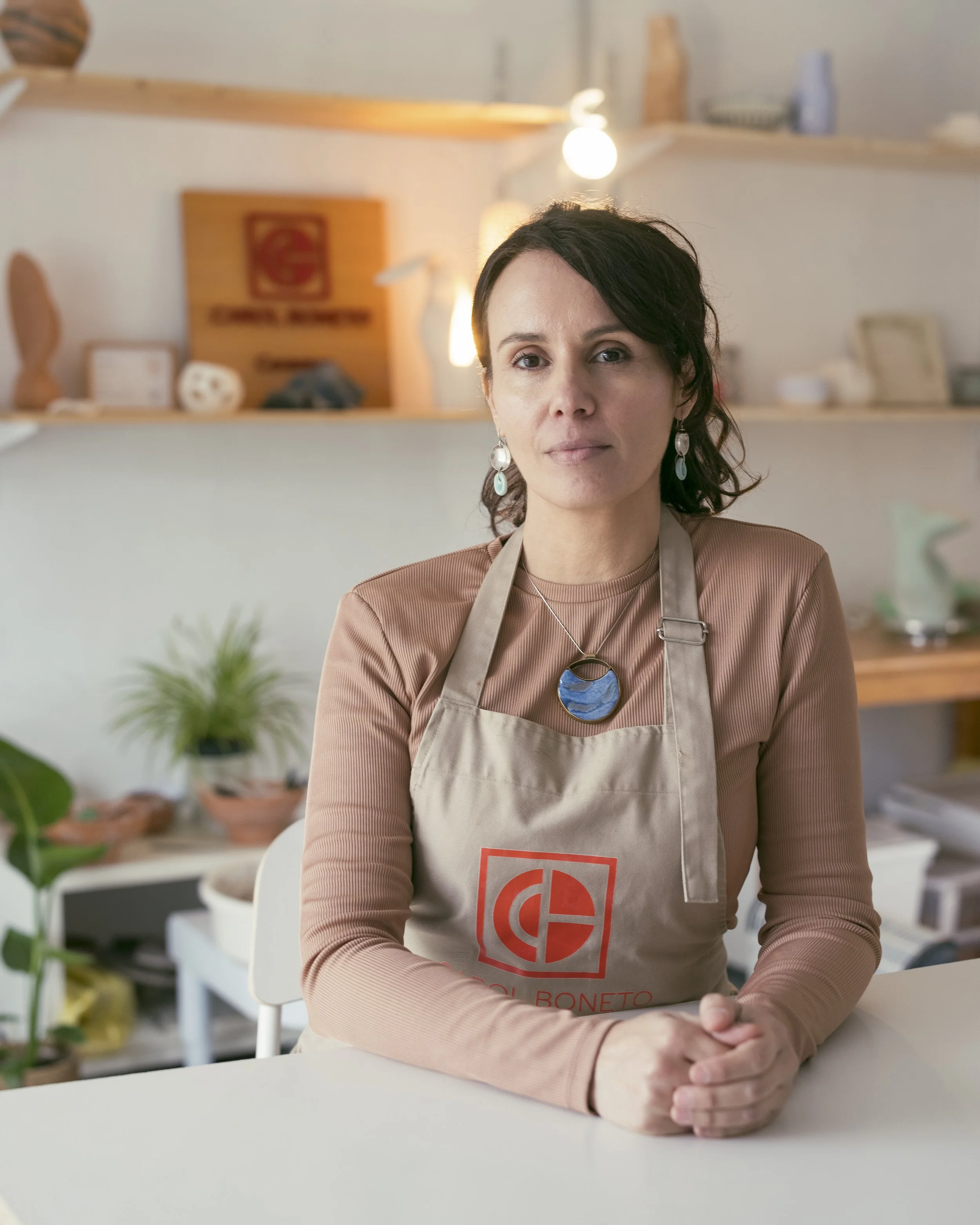 A woman with dark hair and earrings sits at a table in a room with shelves and plants in the background, wearing a beige apron with a red logo and a necklace with a blue circular pendant.