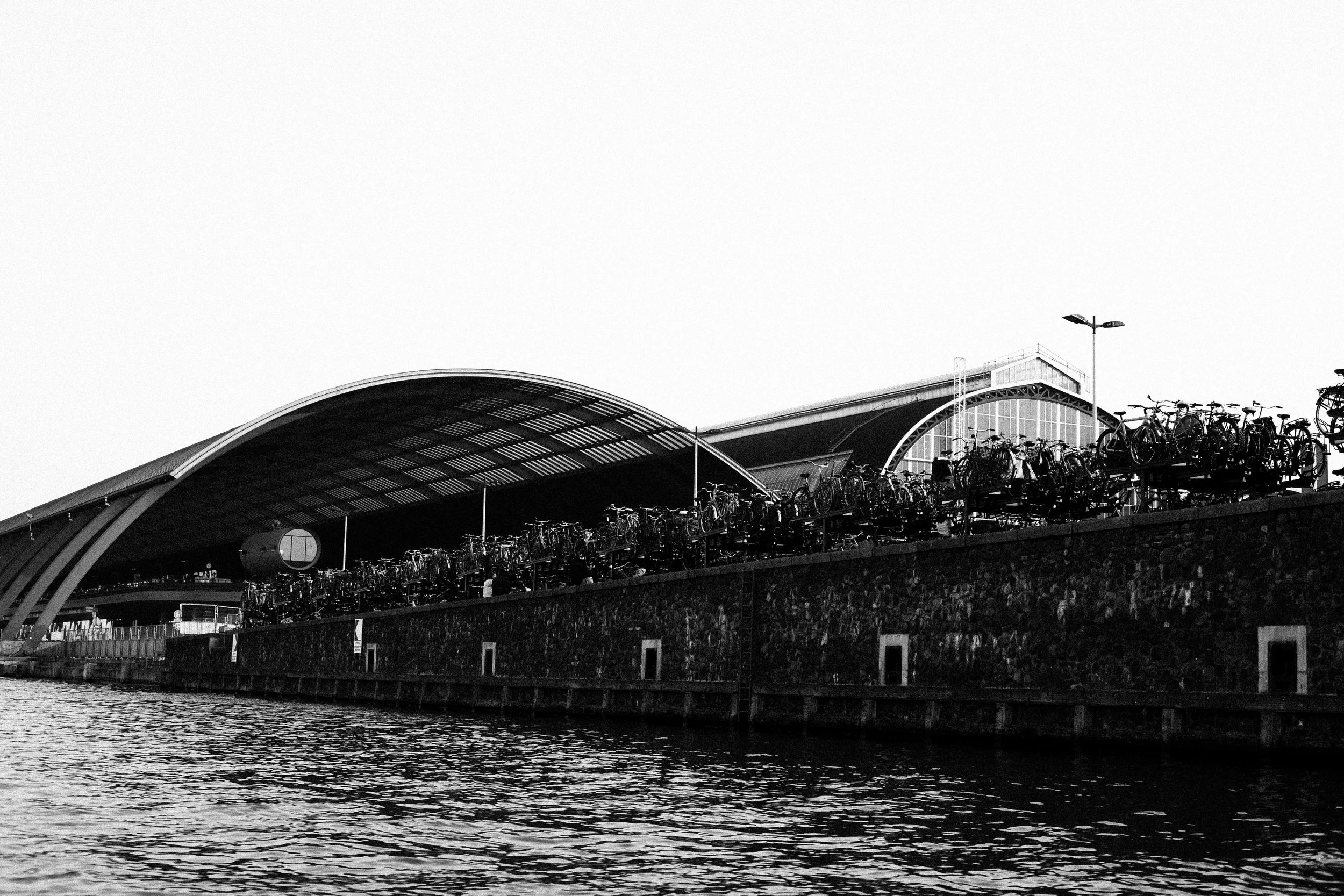 Black and white photo of a modern boat terminal or station with a curved roof, bicycles parked on the rooftop, and water in the foreground.