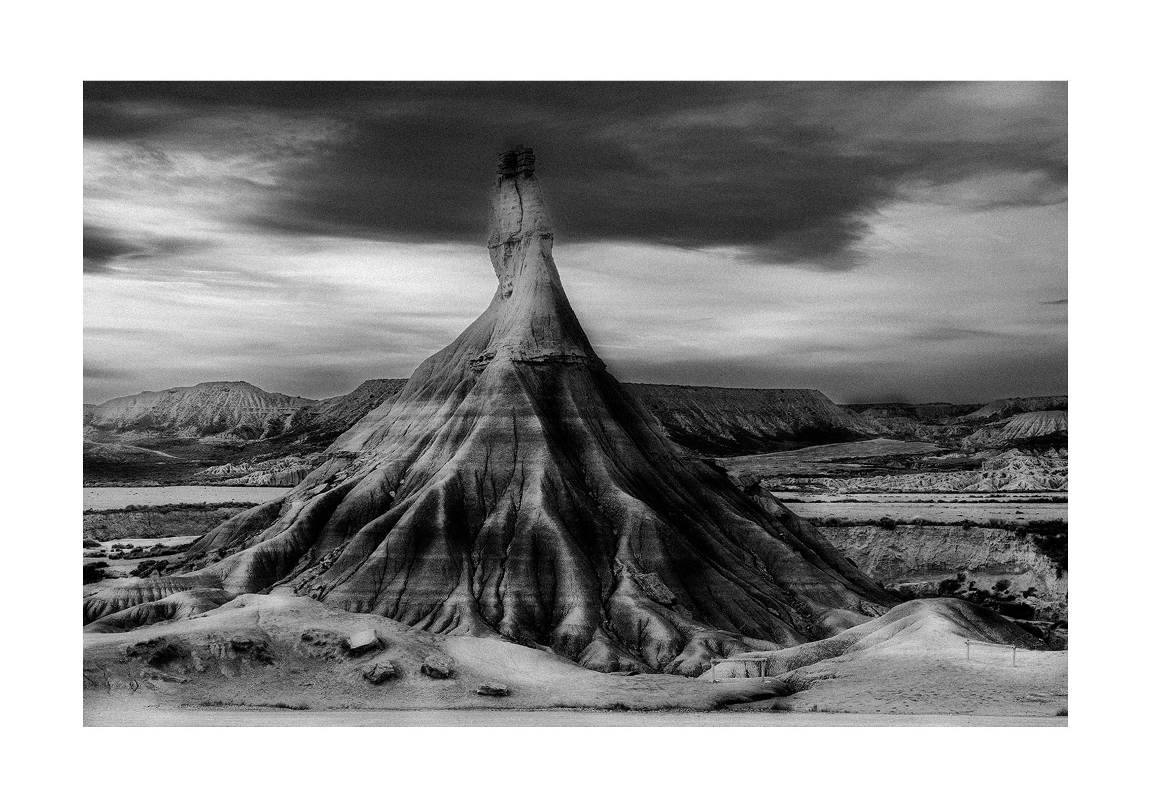 Black and white photo of a tall, cone-shaped mountain with vertical striations, under a cloudy sky, in a desert landscape.