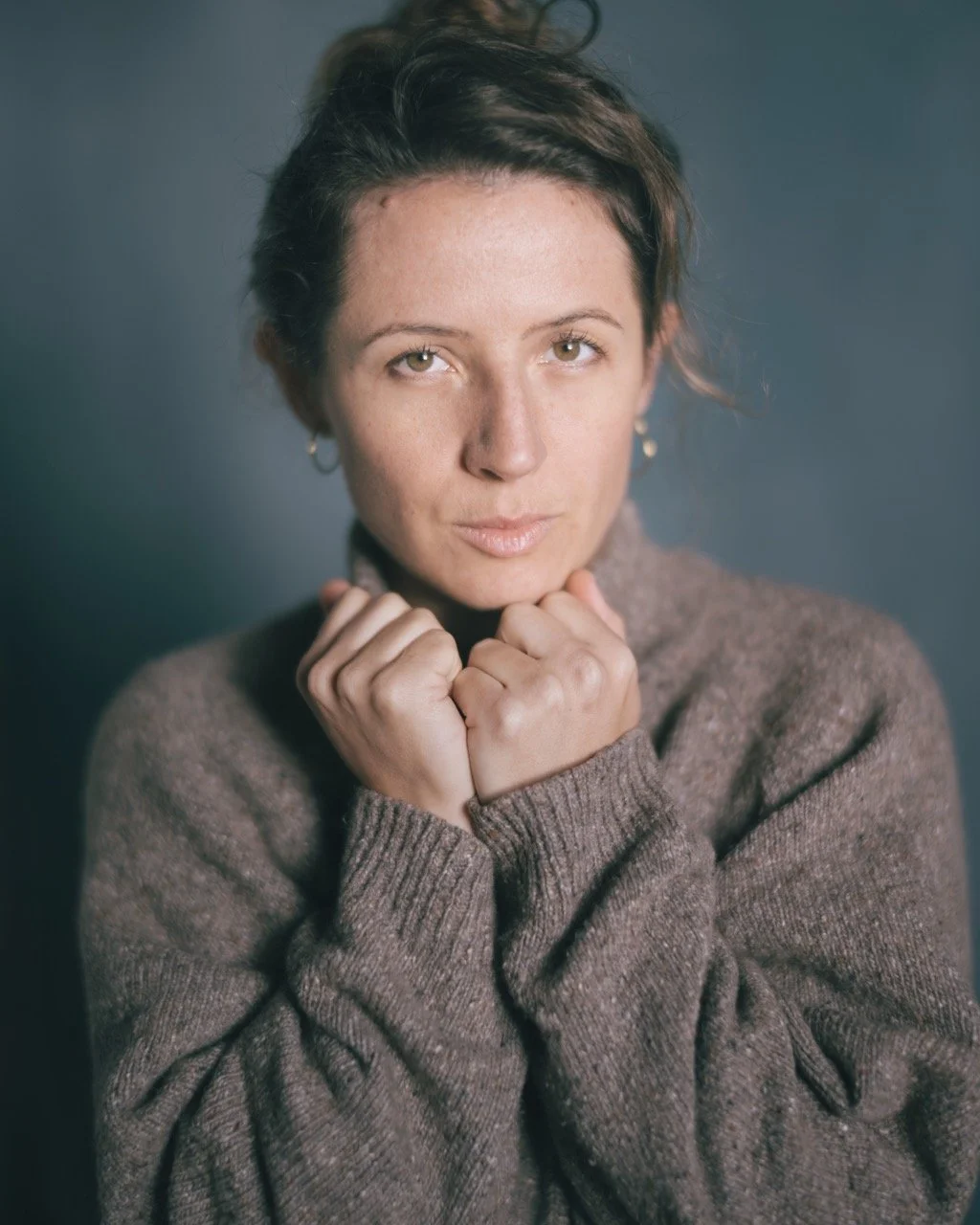 A woman with short brown hair and light skin looks directly at the camera, resting her chin on her clasped hands, dressed in a brown sweater with a soft gray background.