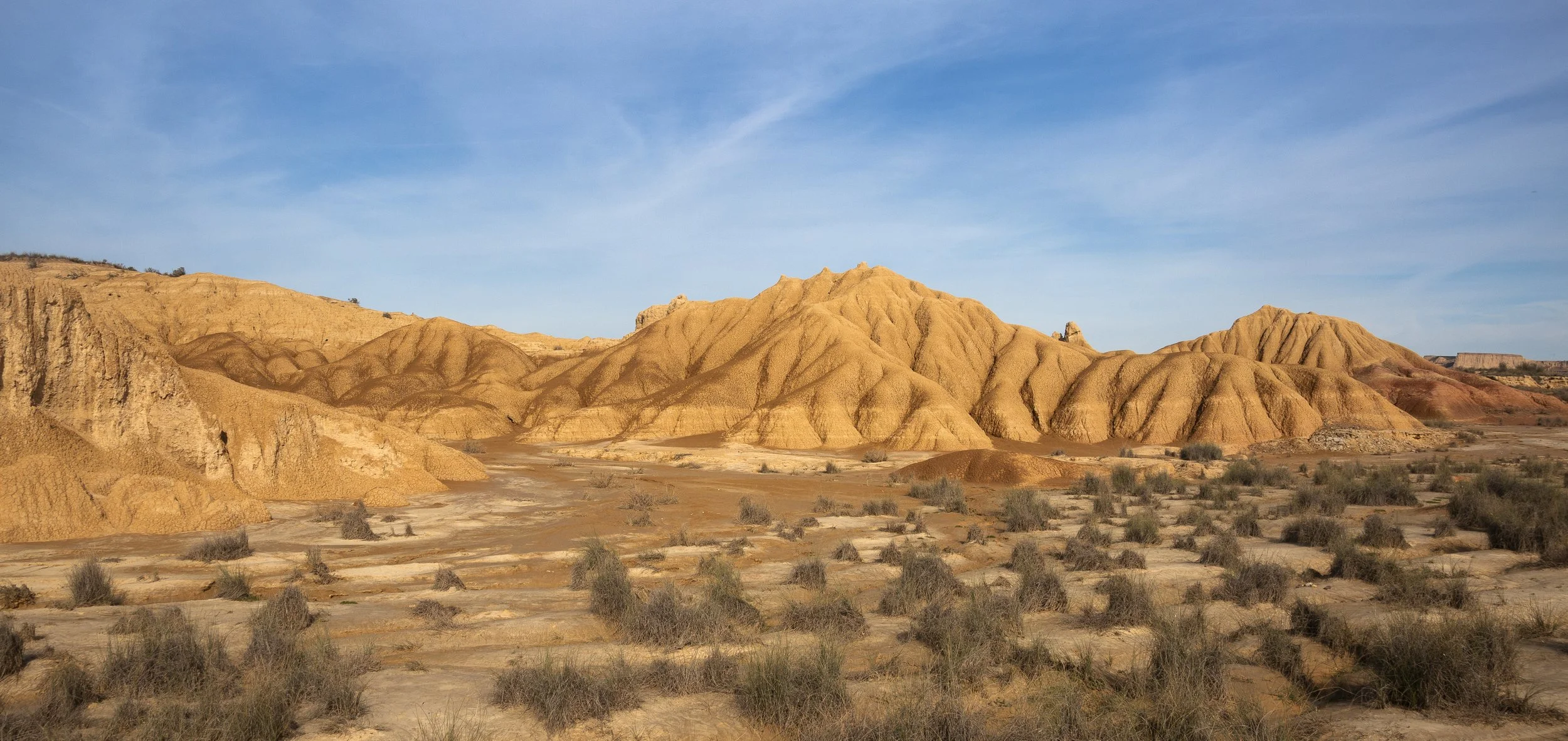 Image of a desert landscape IN THE BARDENAS REALES DESERT IN NAVARRA, SPAIN with orange and yellow rock formations under a clear blue sky, sparse desert shrubs scattered across the ground.