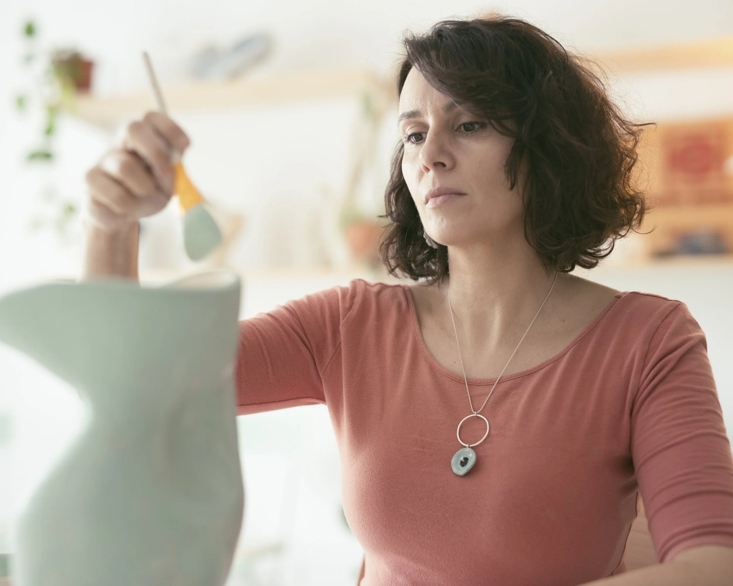 A woman with curly dark hair wearing a salmon-colored top and a necklace with a circular pendant, is painting a ceramic object in a well-lit room.