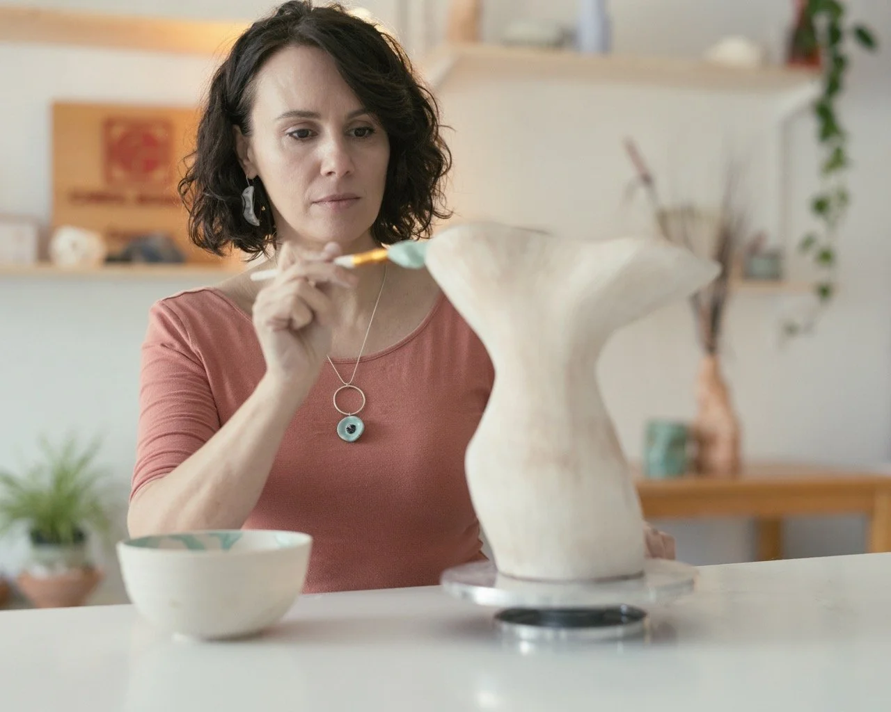 A woman painting a ceramic sculpture with a paintbrush in a brightly lit art studio.