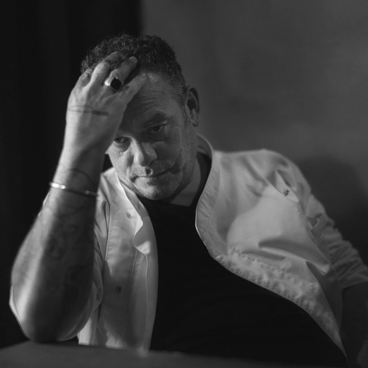 A black and white photo of a man with short, curly hair, wearing a white chef's coat and a black shirt, resting his head on his hand with a contemplative expression.