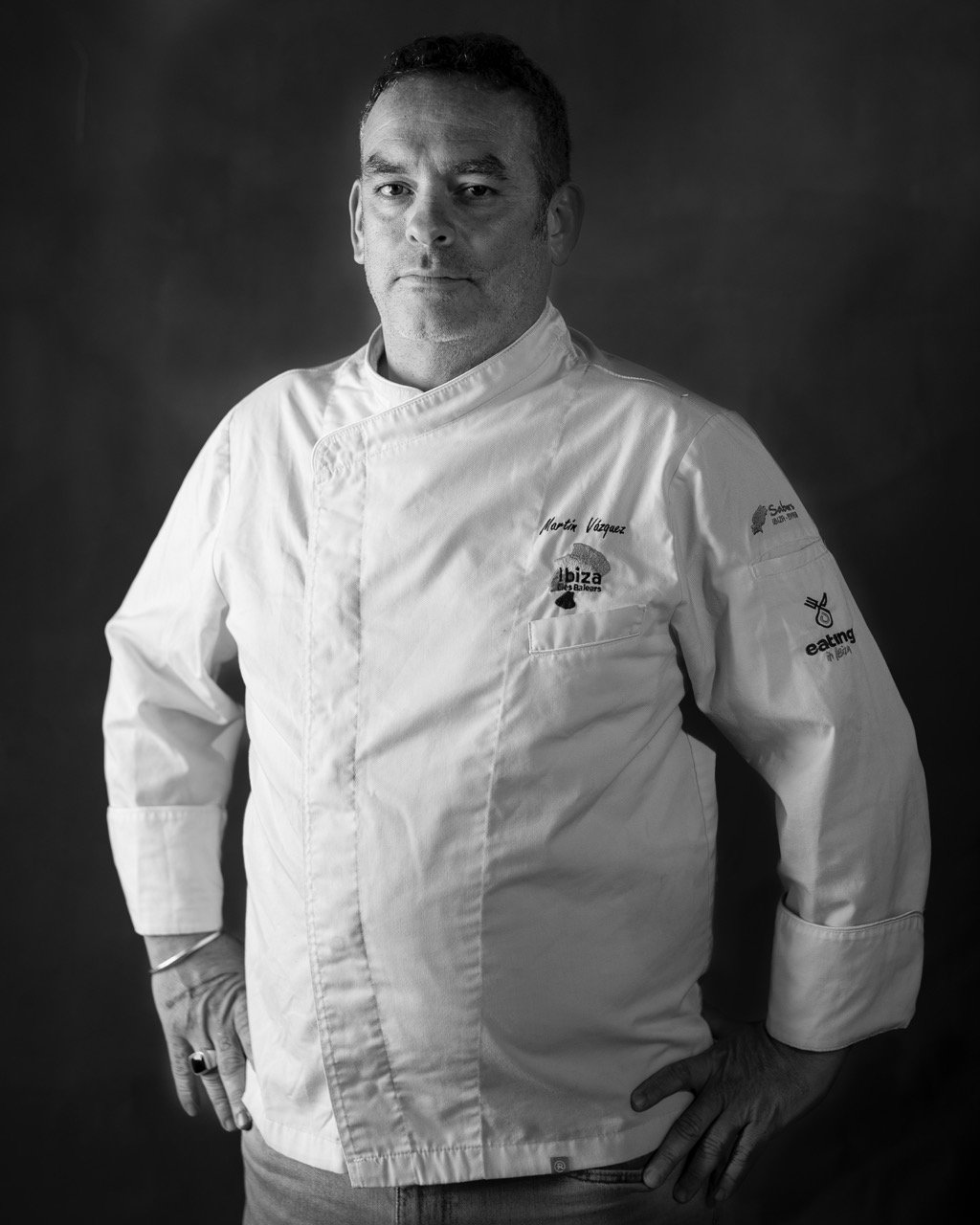 Black and white photo of a male chef in a chef's coat with text and logos, standing with hands on hips, looking at the camera against a dark background.
