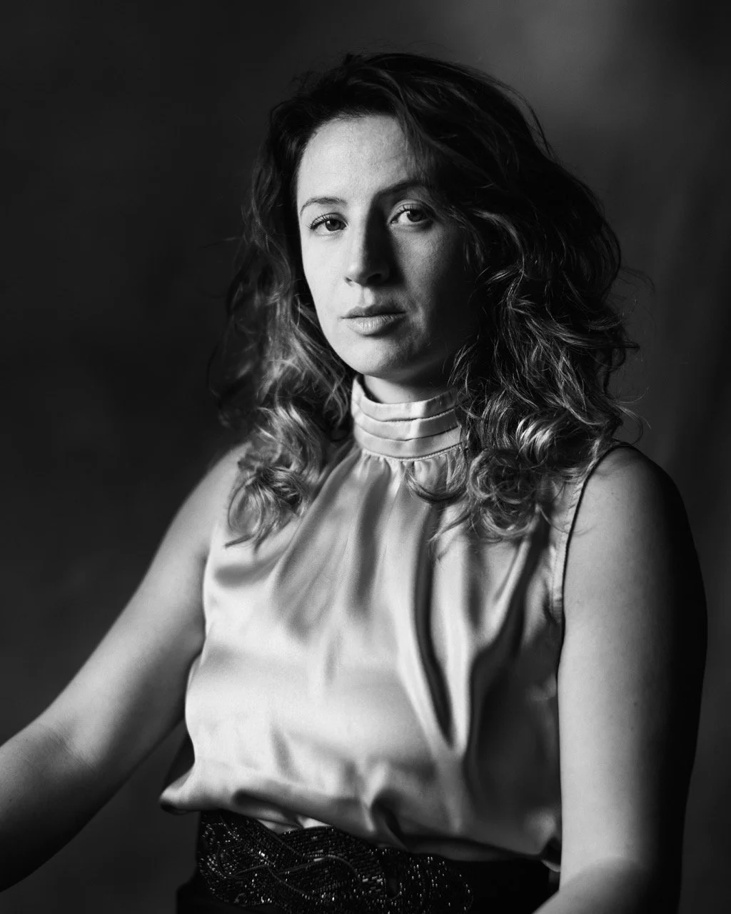 Black-and-white portrait of a woman with curly hair, wearing a high-neck satin top and a skirt, looking at the camera.