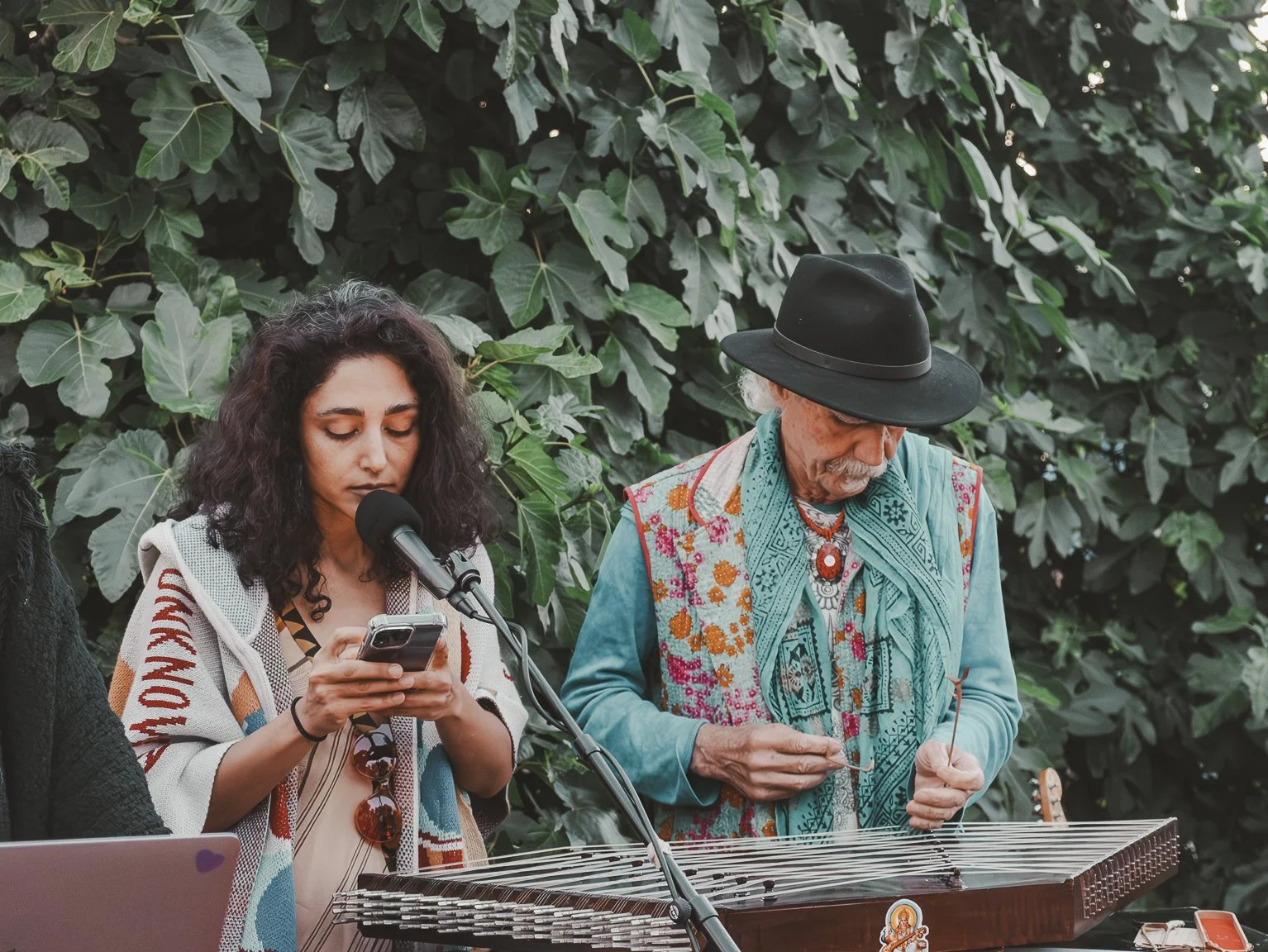 A woman with curly dark hair looking at her phone and a man with gray hair and beard playing a steel guitar outdoors in front of lush green leafy background.