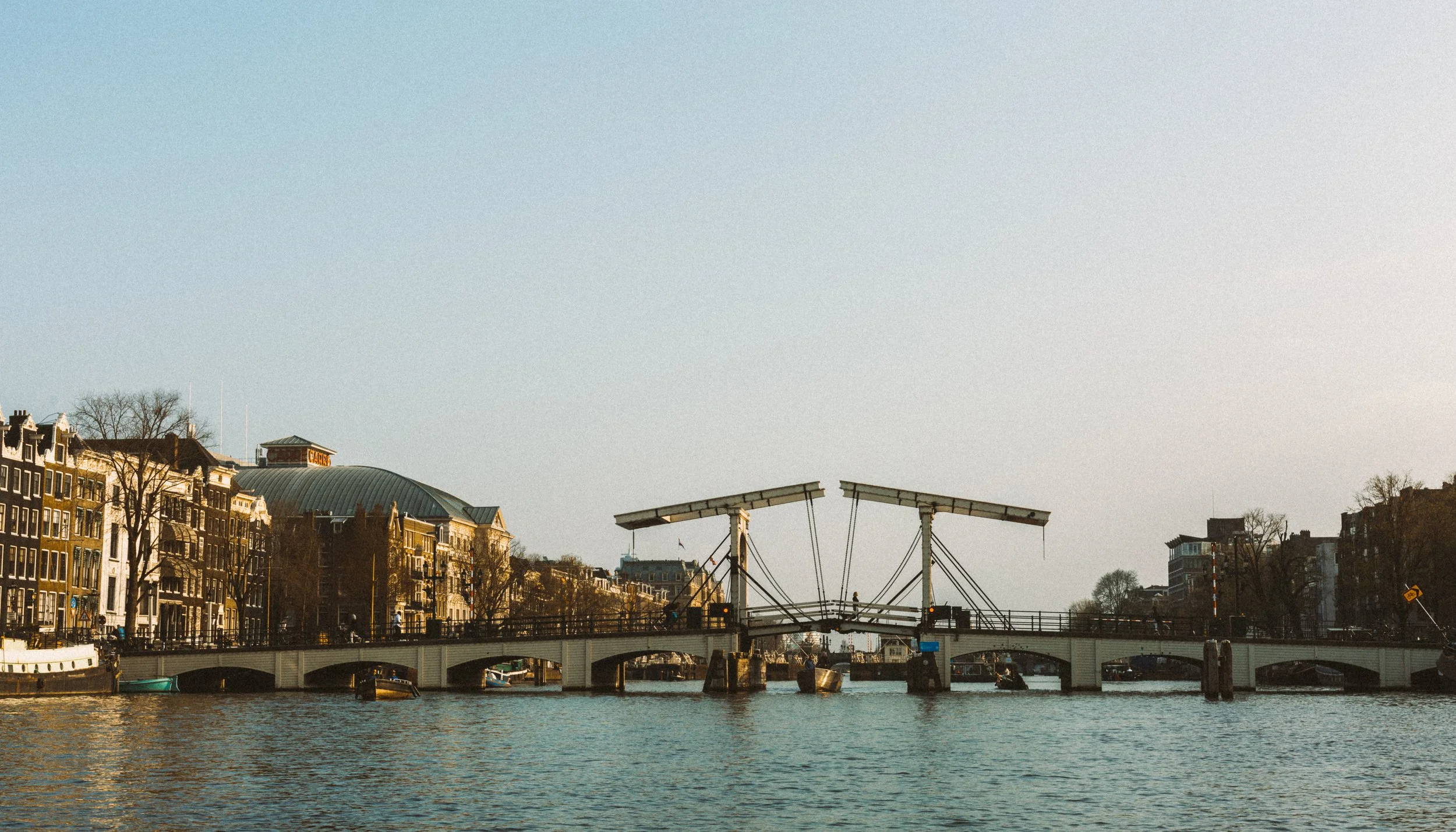 View of a canal with a traditional Dutch water gate lift bridge, surrounded by classic European style buildings along the waterfront, under a clear sky.