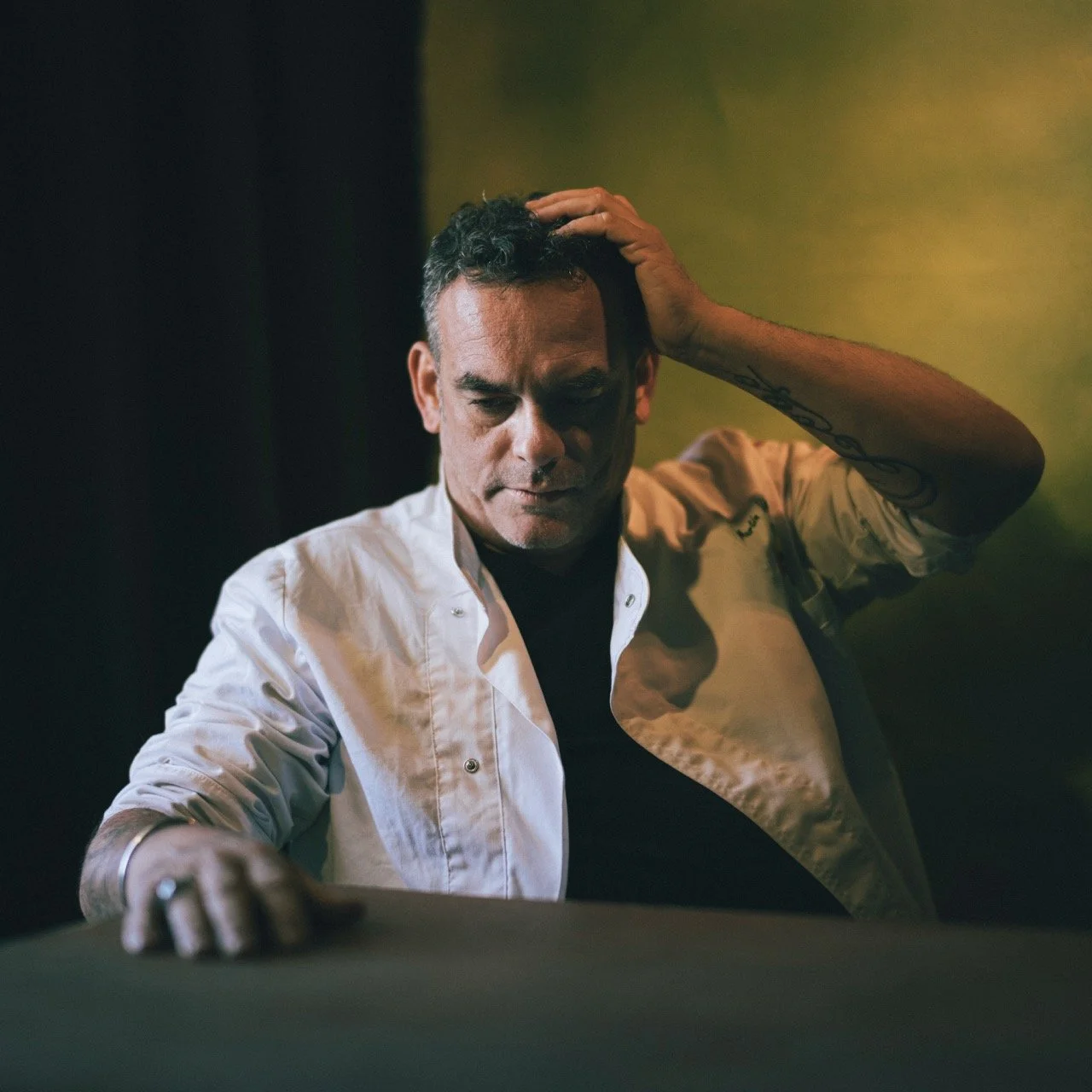 A man with dark hair and tattoos, wearing a white shirt over a black t-shirt, is sitting with a contemplative expression, touching his head with one hand, in a dimly lit room with yellow-green walls.