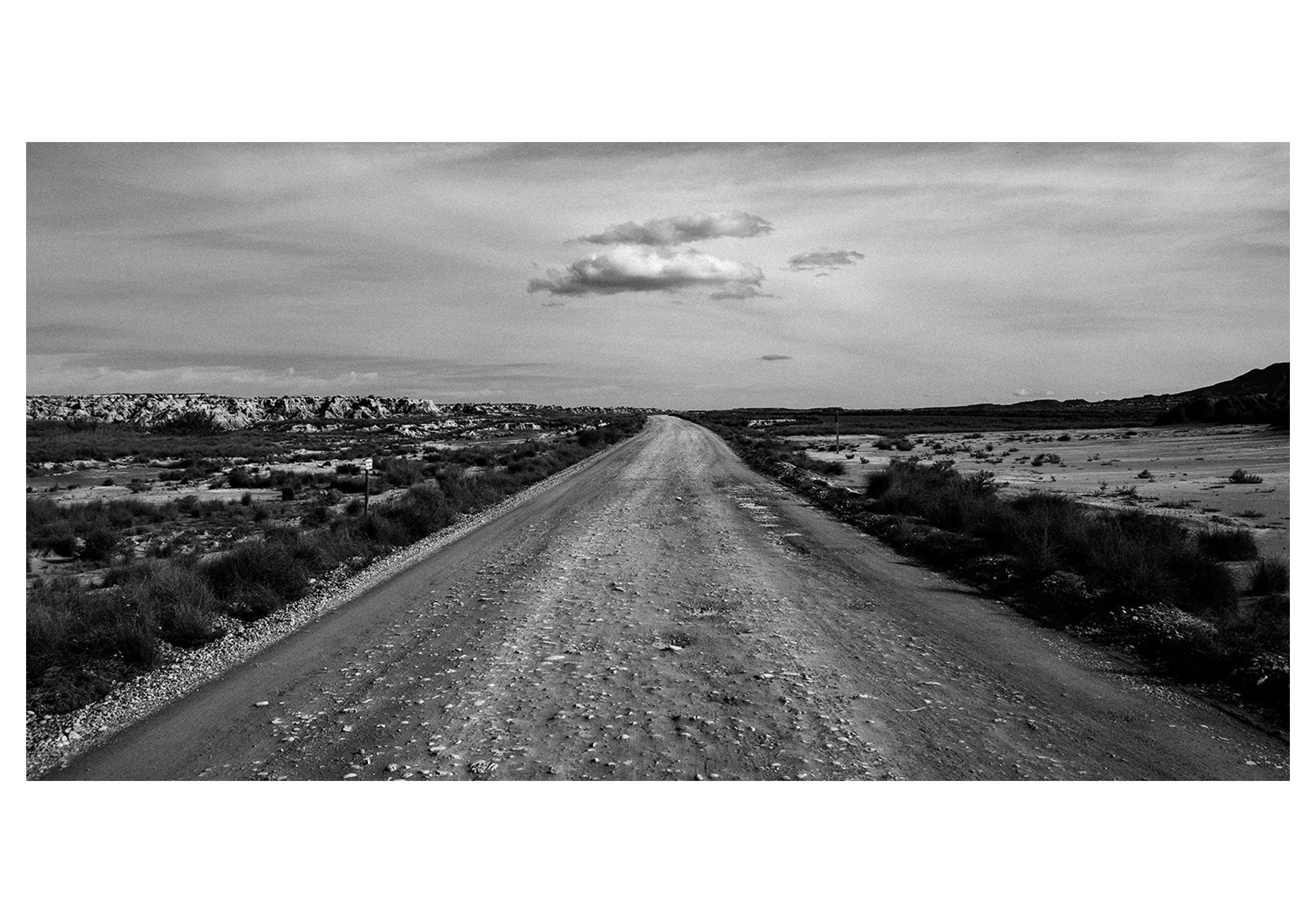 A black and white photo of a dirt road stretching into the horizon through a desert landscape with sparse bushes and mountains in the distance, under a partly cloudy sky.