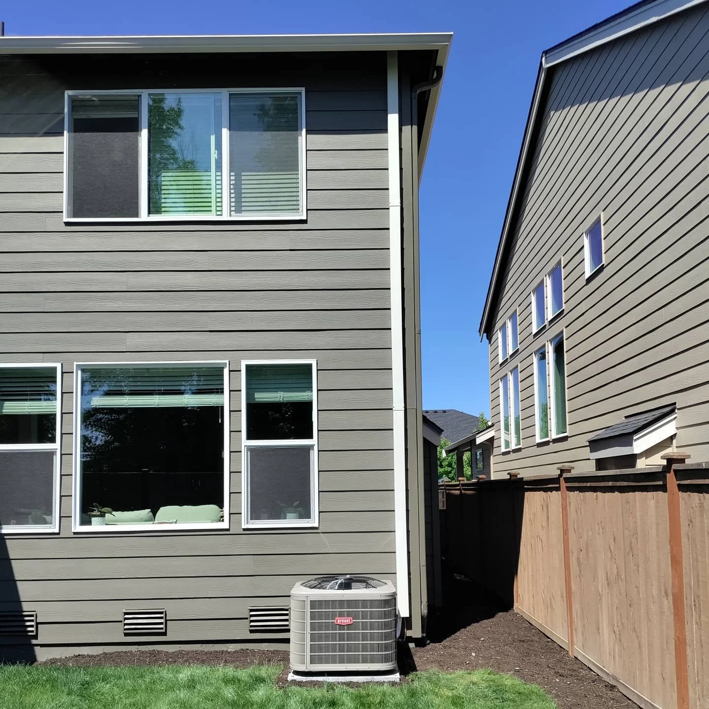 Side view of two modern gray houses with multiple windows, a wooden fence, and an outdoor air conditioning unit in the yard, under a clear blue sky.