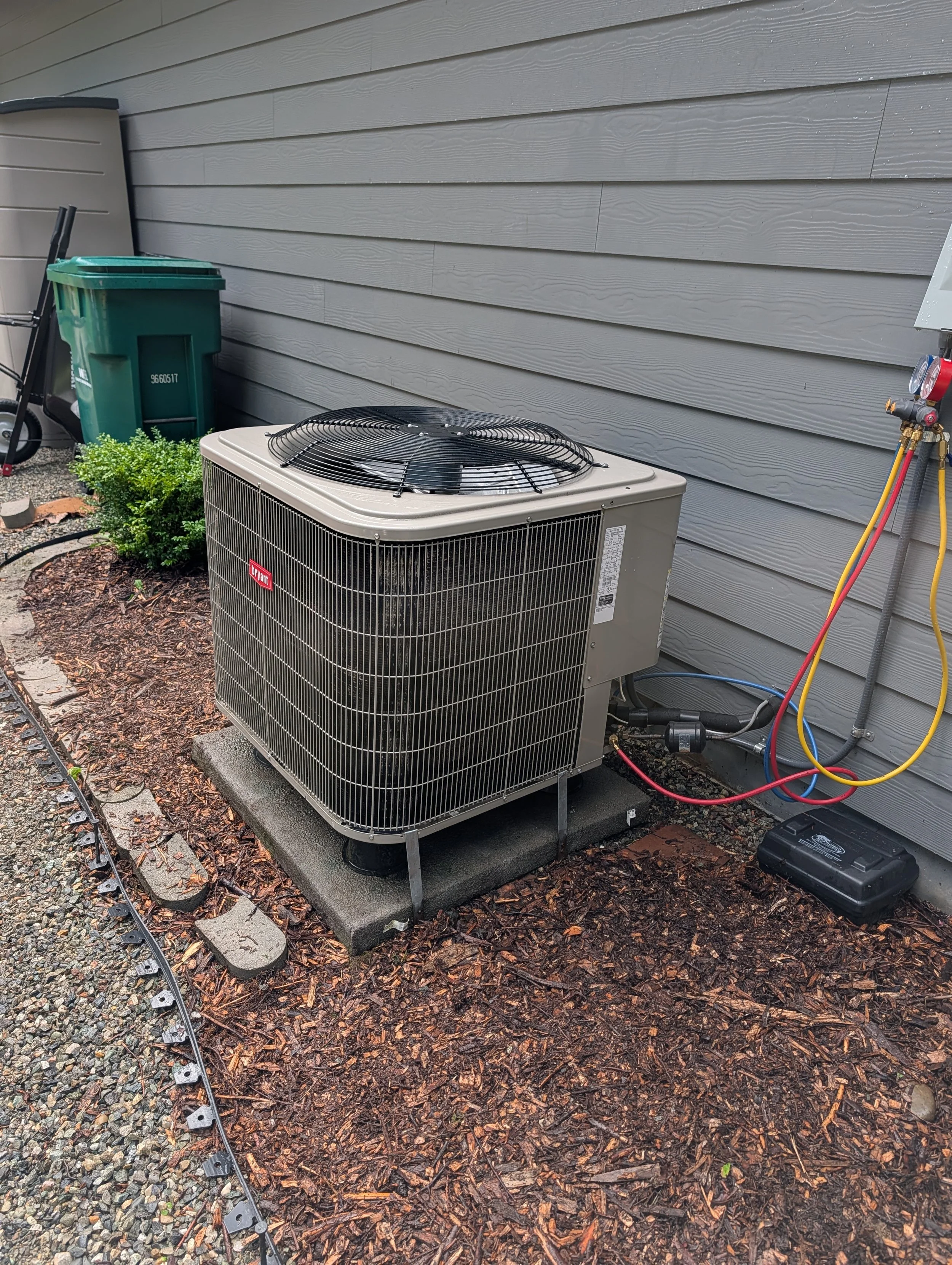 An outdoor air conditioning unit installed on a concrete slab next to a house with gray siding, with electrical connections and hoses nearby.
