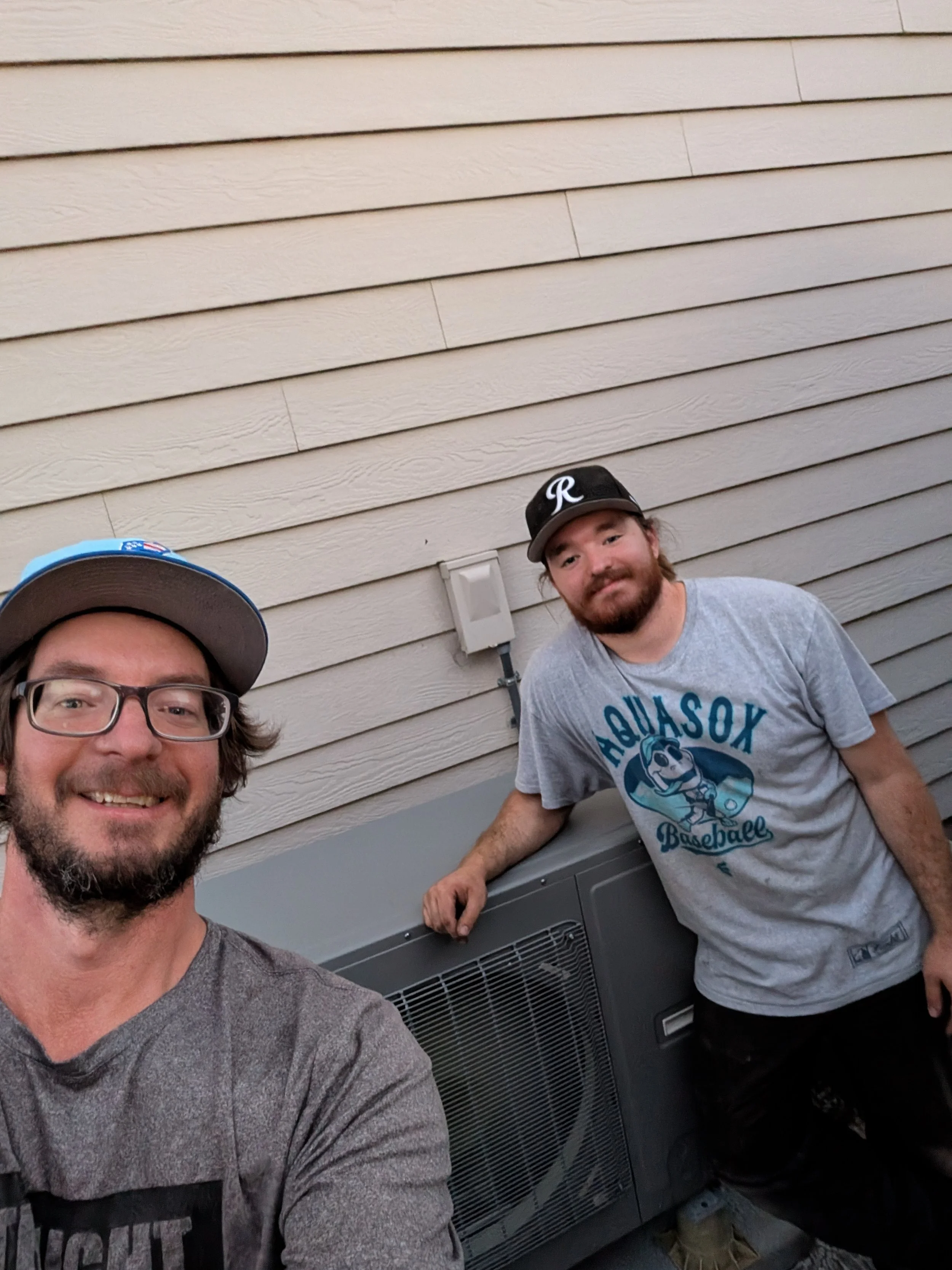 Two men, one with glasses and a cap, smiling and taking a selfie, standing outside near an HVAC unit against a beige siding wall.