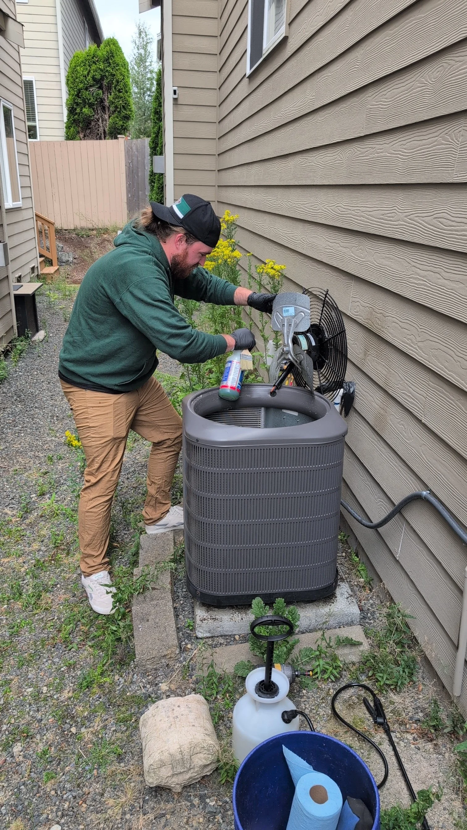 A man wearing a green hoodie, khaki pants, a black cap, and black gloves is cleaning an outdoor air conditioning unit with a spray bottle. He is standing on a gravel path beside a house with beige siding, with gardening plants nearby and a fence in the background.