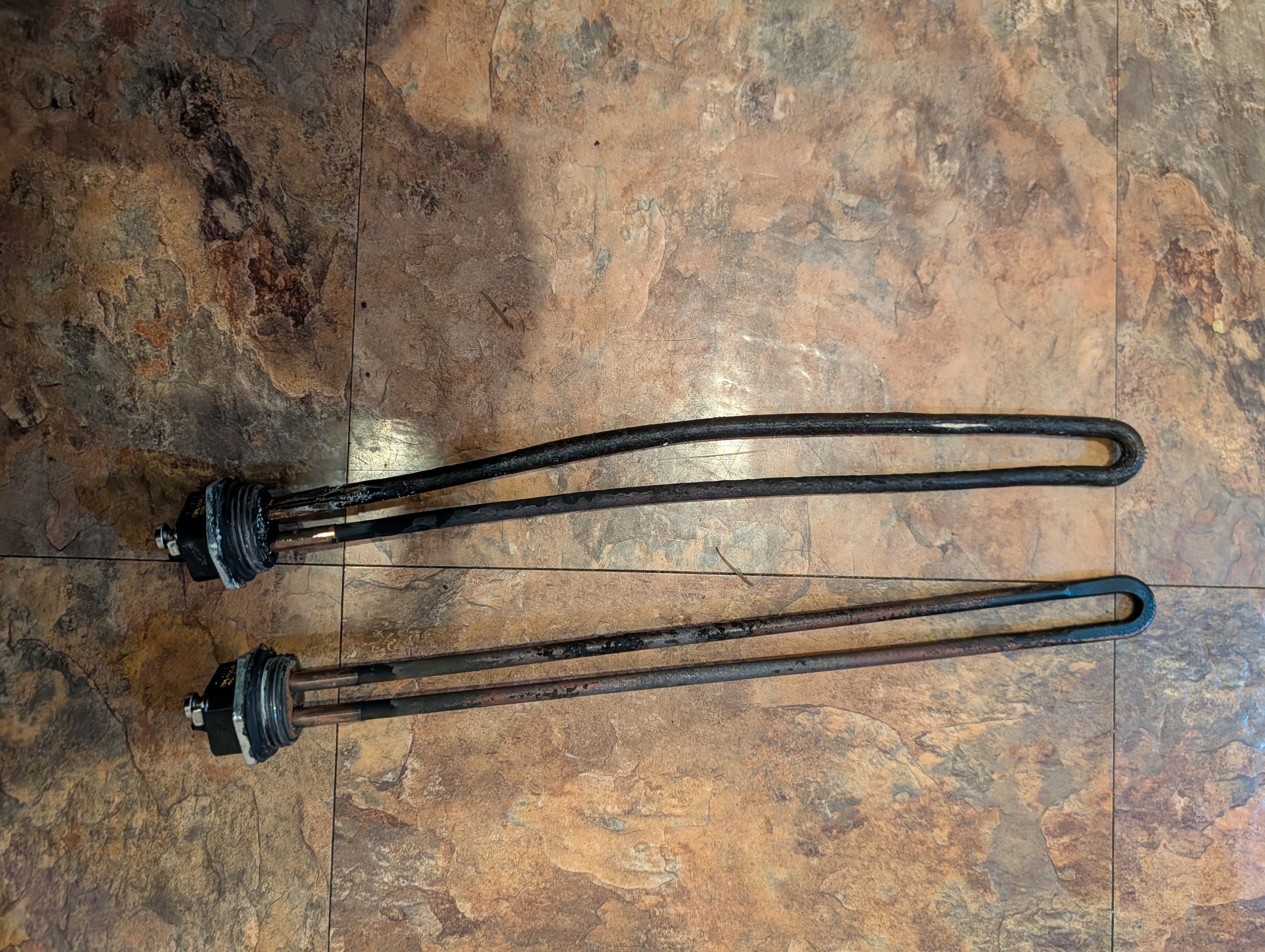 Two metal heating element rods, one with blackened and corroded surface, and the other with rust, resting on a tiled floor.