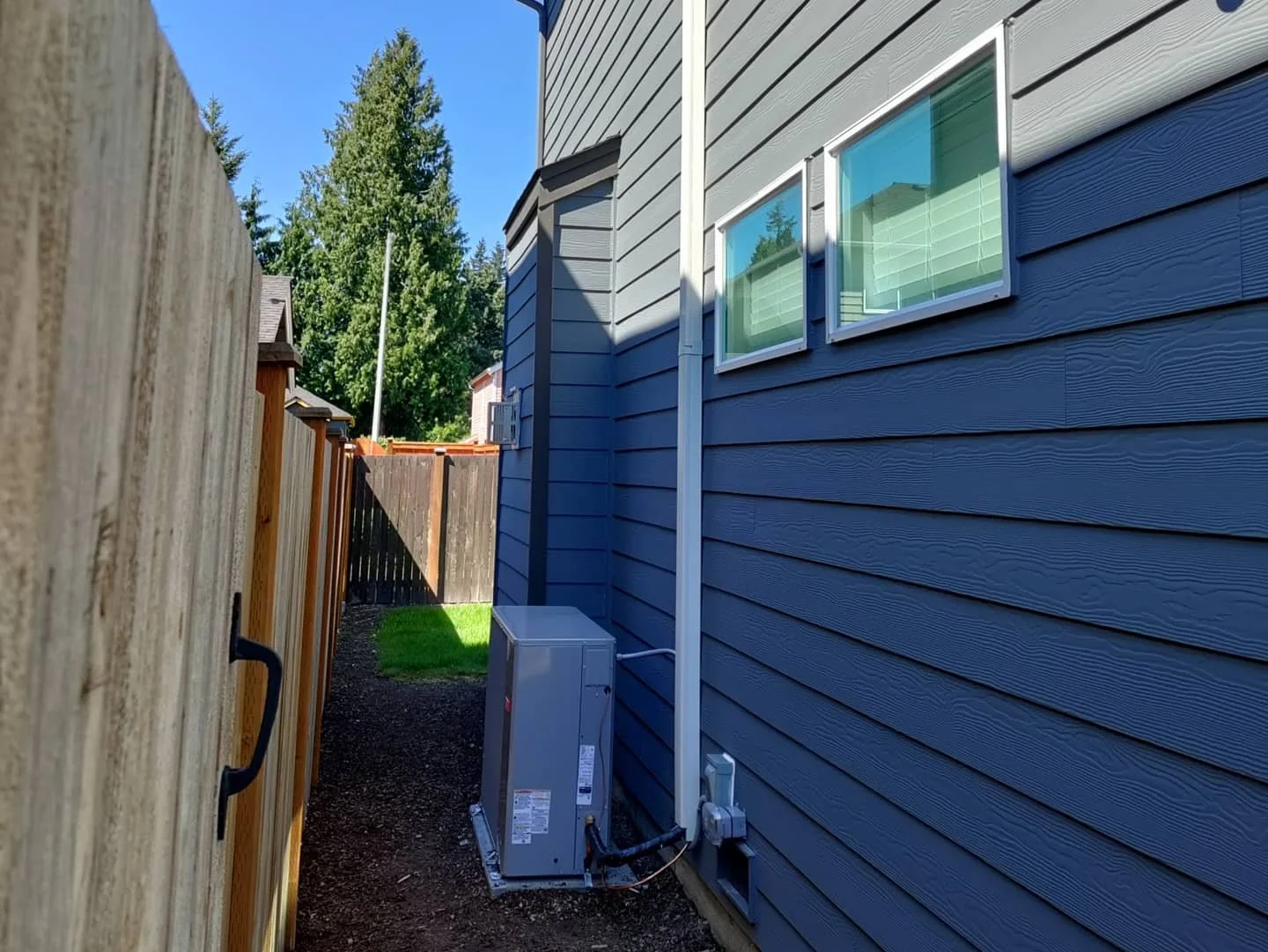 Side view of a blue house with siding, two windows, and an outdoor air conditioning unit on the ground, with a wooden fence on the left and trees in the background under a clear blue sky.